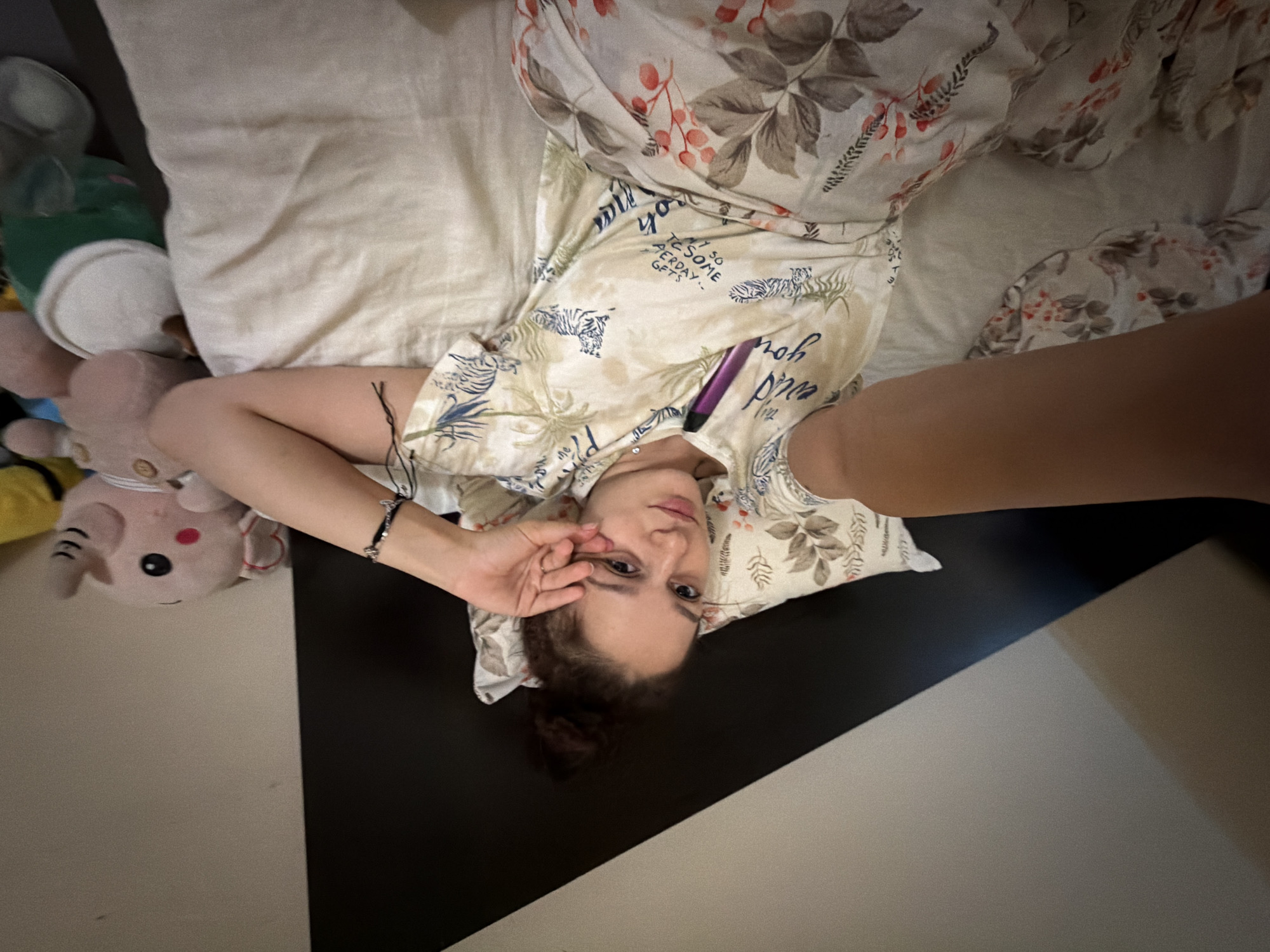 A young woman with her hair in a bun lies on a bed. She is wearing a light-colored t-shirt with a tropical pattern and tigers on it, along with matching pajama bottoms. She is holding her hand up to her face, covering one eye. A pink elephant stuffed animal and a green cup are visible on the bed next to her. The background consists of a diagonally divided wall, with the top section being white and the bottom section black. The scene is indoors, likely a bedroom. There are no discernible landmarks or clues within the image that would indicate its location in Sofia, Bulgaria.