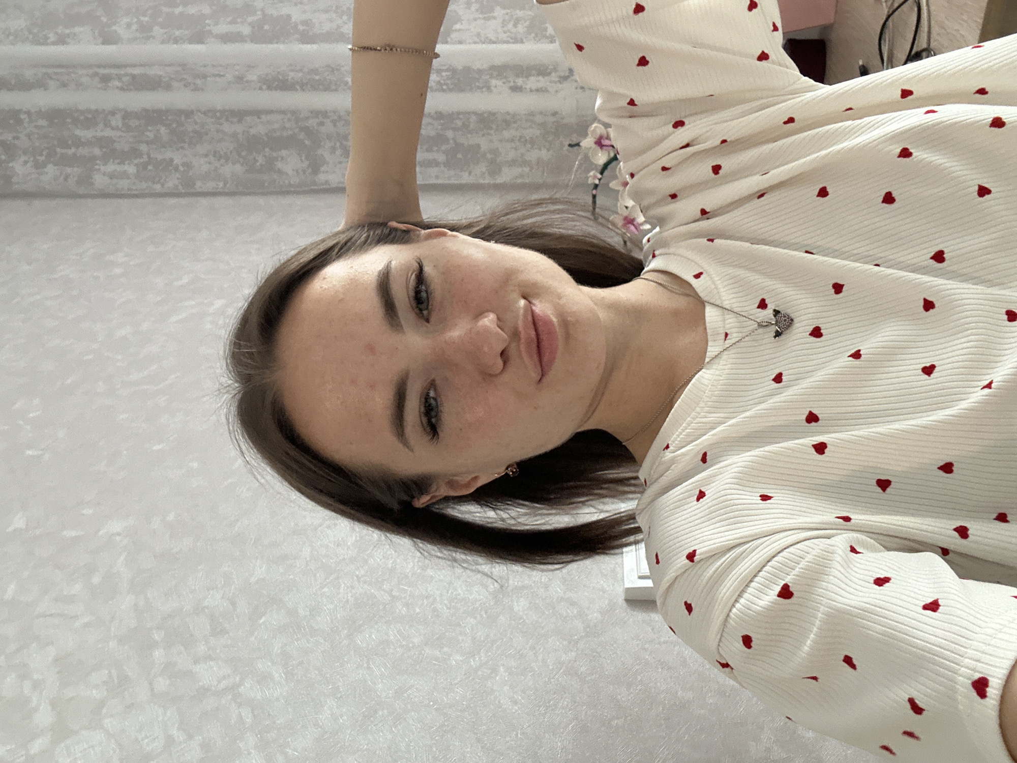 A young woman with brown hair and fair skin with freckles looks at the camera, her right arm bent behind her head. She is wearing a white t-shirt with red heart patterns and a silver necklace. The background appears to be a room with textured wallpaper and curtains.