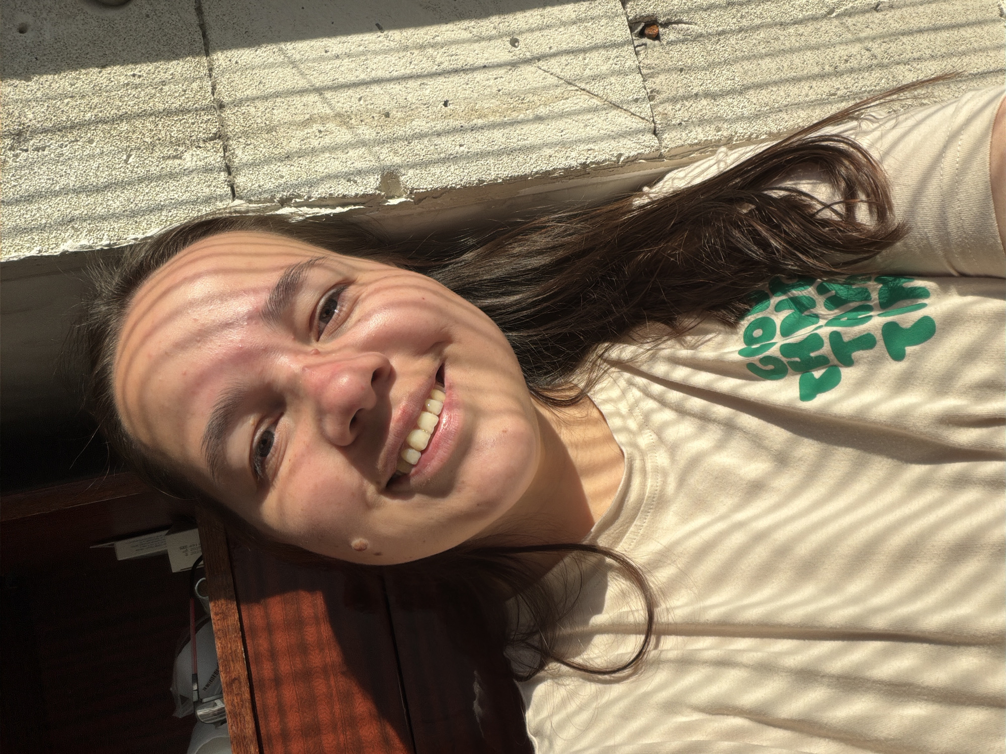 A young woman is lying down, smiling at the camera. She is wearing a light beige t-shirt with a green logo on the upper left side. Her dark brown hair is spread out around her head. Light and shadows from a window blind or similar structure fall across her face and t-shirt. Her head rests on a dark wooden surface, possibly a desk or table. The backdrop consists of a grey brick wall. A city or location is not explicitly identifiable in the image.