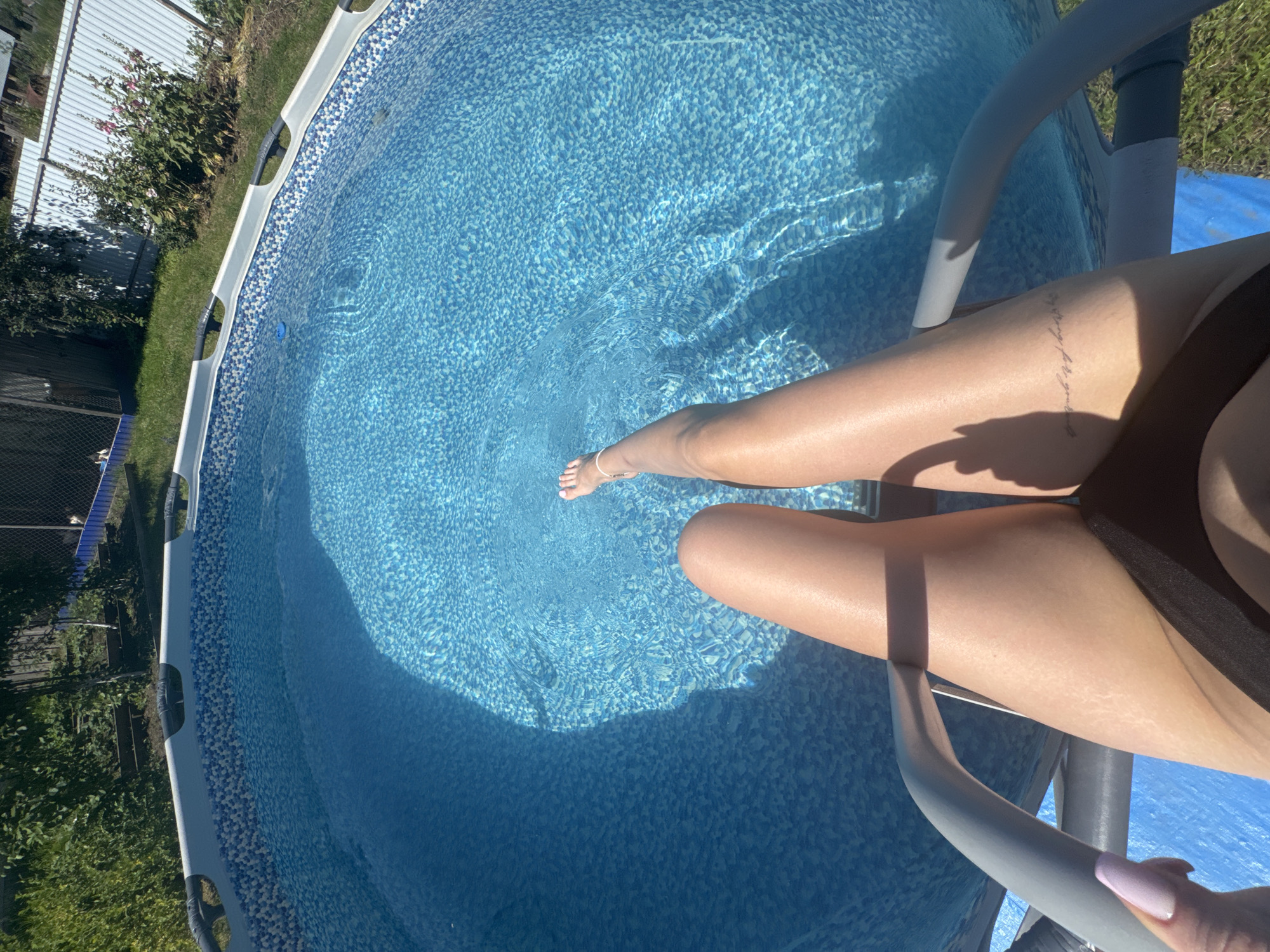 In the city of Olkhovka, Russia, a person's legs and feet are submerged in a blue, patterned above-ground swimming pool. The person is wearing a black bikini bottom and has a delicate anklet on their left ankle. A tattoo is visible on their right thigh. The pool is situated outdoors, surrounded by green grass, and a grey metal ladder is partially visible in the foreground. In the background, a white fence and lush green foliage can be seen. The water is clear, with ripples and light reflections indicating sunlight.