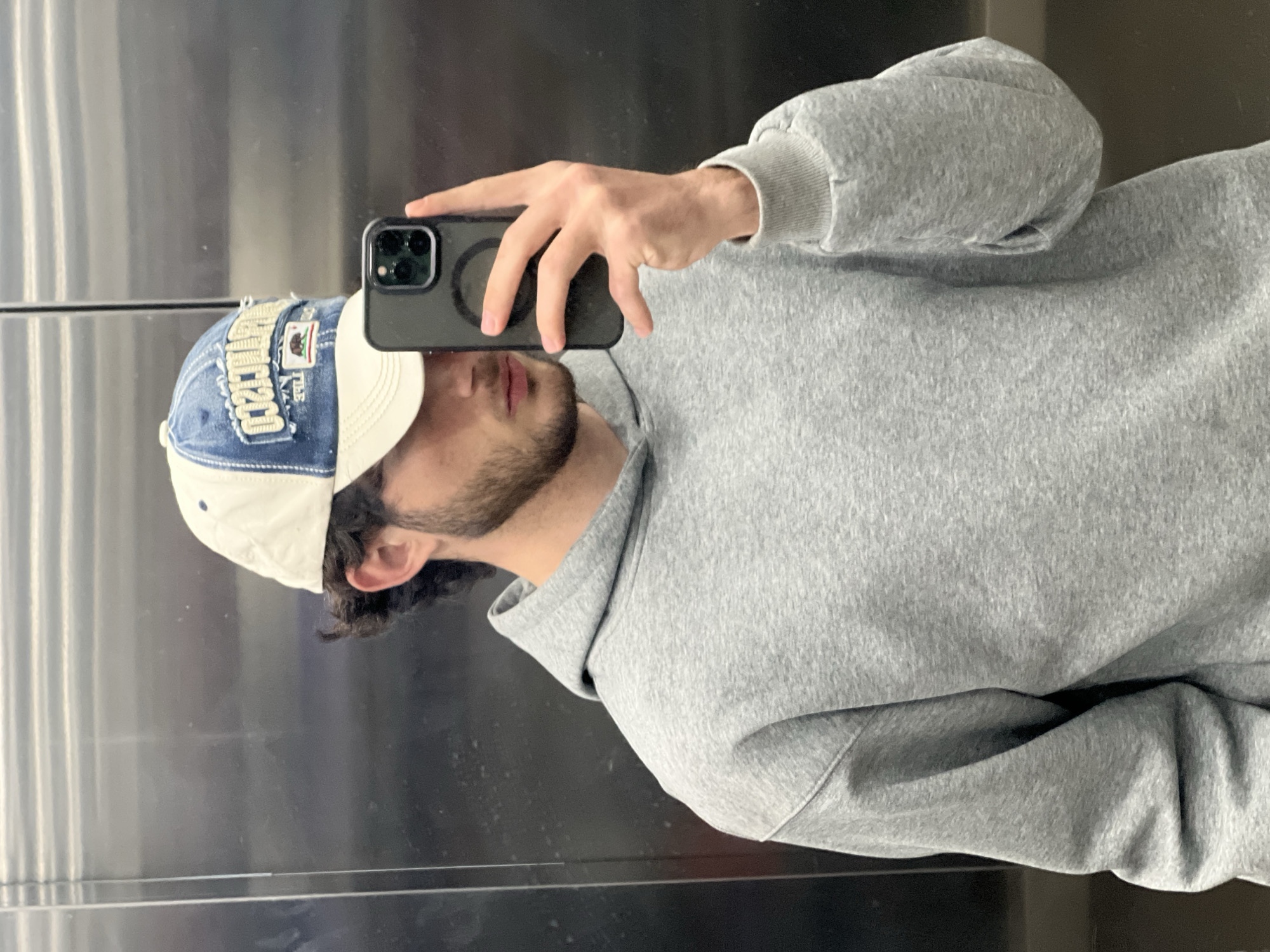 A man is taking a selfie in front of a reflective surface, possibly an elevator. He is wearing a grey hoodie and a two-tone baseball cap with a denim-like upper section and a cream-colored brim. He holds a black smartphone in his hand, obscuring part of his face. The background is metallic and featureless. The image location is unspecified.