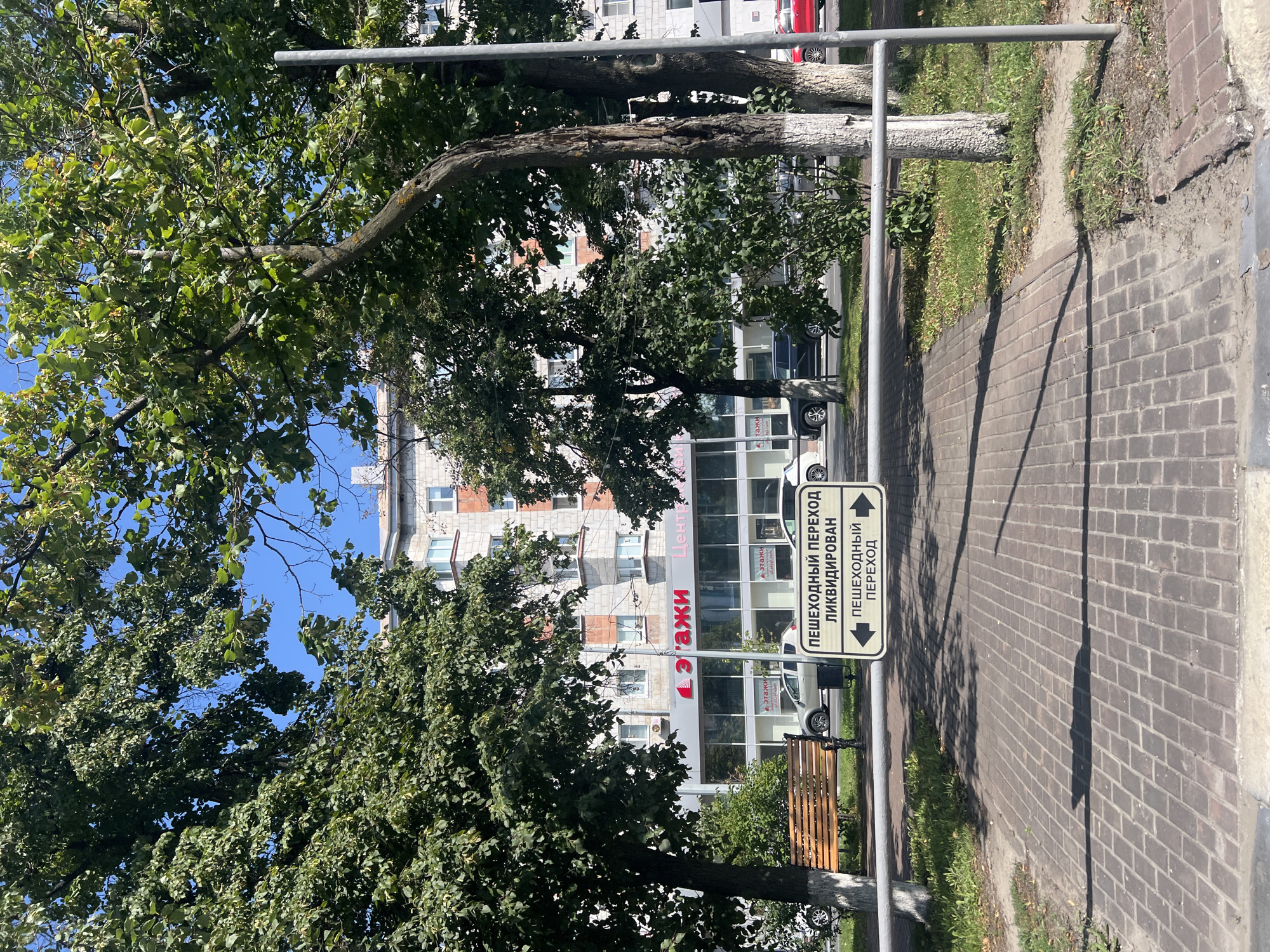 A horizontal, metallic barrier is positioned across a brick-paved walkway. Mounted on this barrier is a white sign with black text and directional arrows. The top line of text reads "ПЕШЕХОДНЫЙ ПЕРЕХОД" (Pedestrian Crossing), followed by "ЛИКВИДИРОВАН" (Liquidated/Removed) on the second line. The third line of text is also "ПЕШЕХОДНЫЙ ПЕРЕХОД", with a left-pointing arrow on the left and a right-pointing arrow on the right.
Behind the barrier, a grassy area with scattered trees is visible. In the background, a multi-story building with a light-colored brick facade and visible windows stands under a clear blue sky. A light-colored car is parked on the street in front of the building, and a darker colored car is driving away from the camera. The scene appears to be located in an urban environment, likely a residential or commercial area. The signage suggests a change or removal of a designated pedestrian crossing.
Based on the architecture and typical signage found in Russia, this image is likely from Ulyanovsk, Russia. The style of the building and the street furniture are consistent with urban landscapes in Russian cities.