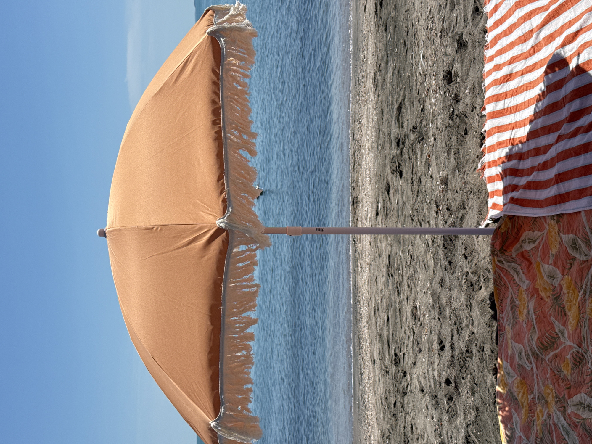 An orange beach umbrella with fringe is centered on a sandy beach under a clear blue sky. The umbrella is casting a shadow on the sand. In front of the umbrella, two beach towels are spread out: one with a tropical leaf pattern in shades of red and yellow, and another with orange and white stripes. The ocean, a deep blue, is visible behind the umbrella. The scene appears to be a serene beach setting, likely in Burgas, Bulgaria, given the context provided. There are no people visible in the frame.