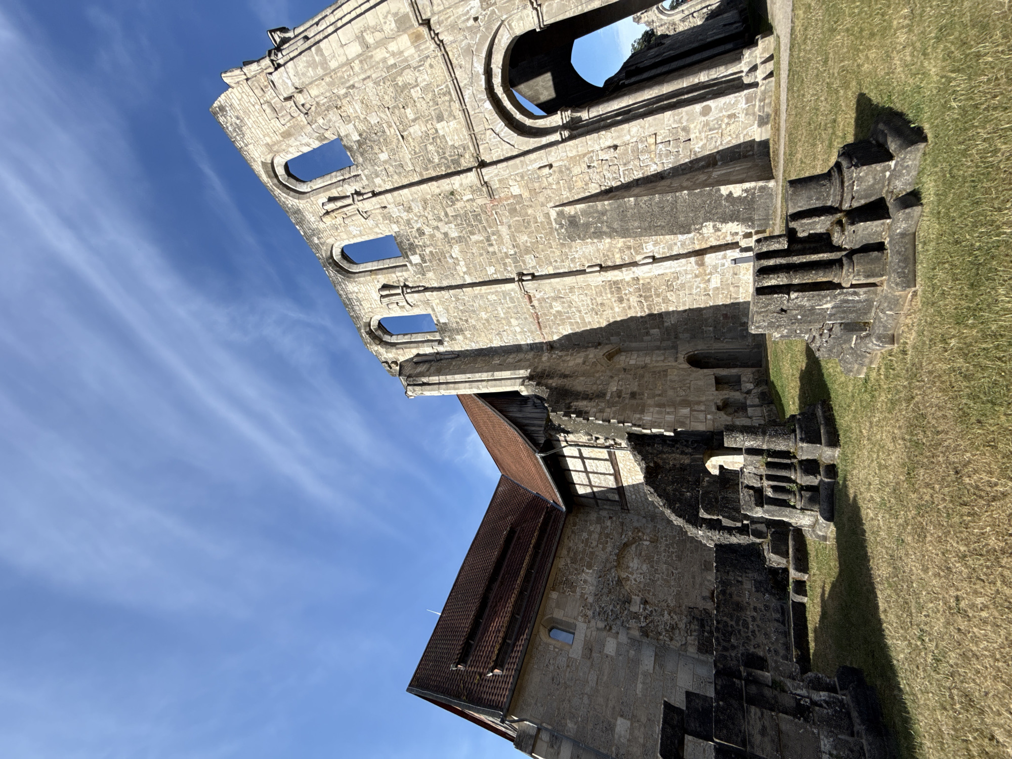 The image shows the ruins of the Walkenried Abbey in Walkenried, Germany. The abbey is made of stone and has a large arched doorway and windows. The sky is blue with some white clouds. In the foreground, there are some stone ruins and grass. The photo is taken from a low angle, looking up at the abbey.