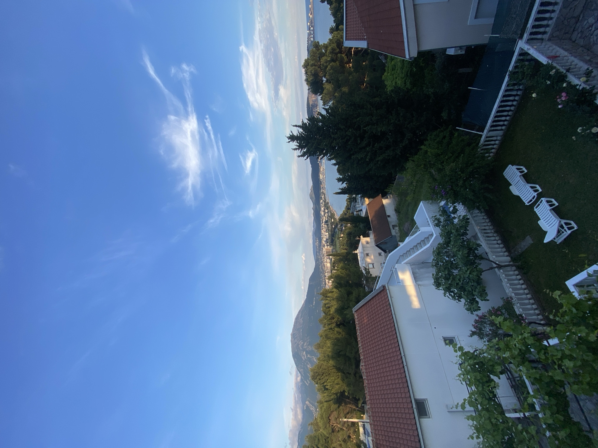 The image captures a partially obscured view of the Adriatic coastline from what appears to be a residential area in Šušanj, Montenegro. A blue sky with wispy clouds fills the upper portion of the frame, juxtaposed against a mountain range covered in lush green vegetation. The camera angle looks down on a residential property featuring white buildings with red-tiled roofs, a manicured lawn, and sun loungers. A body of water (likely the Adriatic Sea) extends into the distance, with distant buildings along the shore, likely a part of the Montenegrin coast.