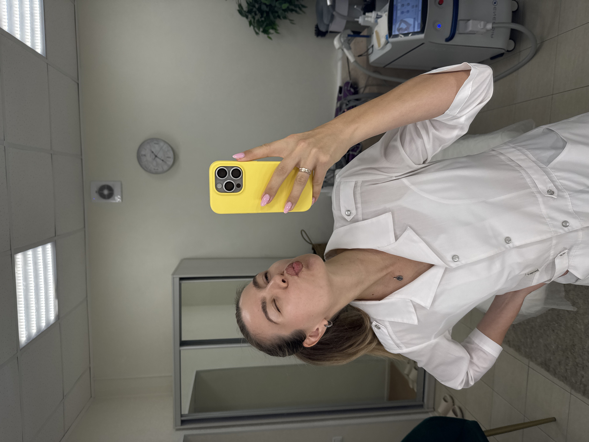 A woman with blonde hair pulled back in a ponytail, wearing a white lab coat and a silver necklace, stands in a room and takes a selfie. She is puckering her lips and winking at the camera, holding up a yellow iPhone in her right hand. Her fingernails are painted pink. A ring is visible on her left ring finger. In the background, a large mirror reflects the room, showing a sliding door wardrobe, a ceiling light, a wall clock, and a piece of medical equipment. The room appears to be a medical or cosmetic office.