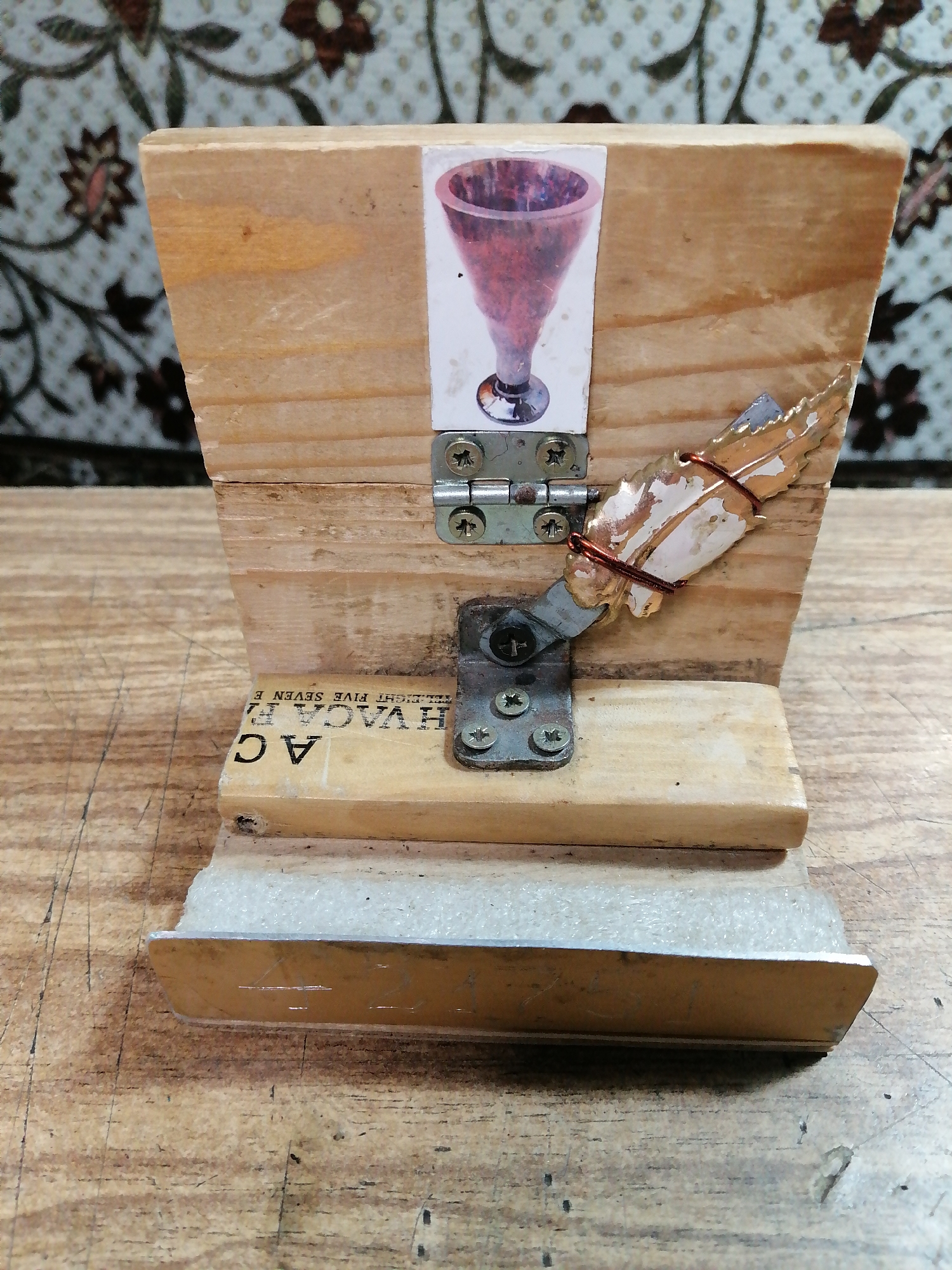 The image shows a handcrafted wooden object, possibly a phone stand, placed on a wooden surface. The object has a tilted rectangular wooden back, with a printed image of a red goblet adhered to its upper portion. A small metal hinge is attached to the back of the stand, with a piece of what appears to be a claw or a similar appendage extending from it, wrapped with copper wire. Below the hinge, a piece of tape with black lettering, partially obscured and reading "HVAC A F", is attached to the front of the stand. The lower part of the stand has a horizontal wooden base with a metallic strip along its front edge. The background is a patterned fabric with floral motifs. The overall scene suggests an indoor setting with natural wood textures dominating the foreground. There is no identifiable location within the city of Rostov-on-Don, Russia, or any specific actions or interactions taking place.