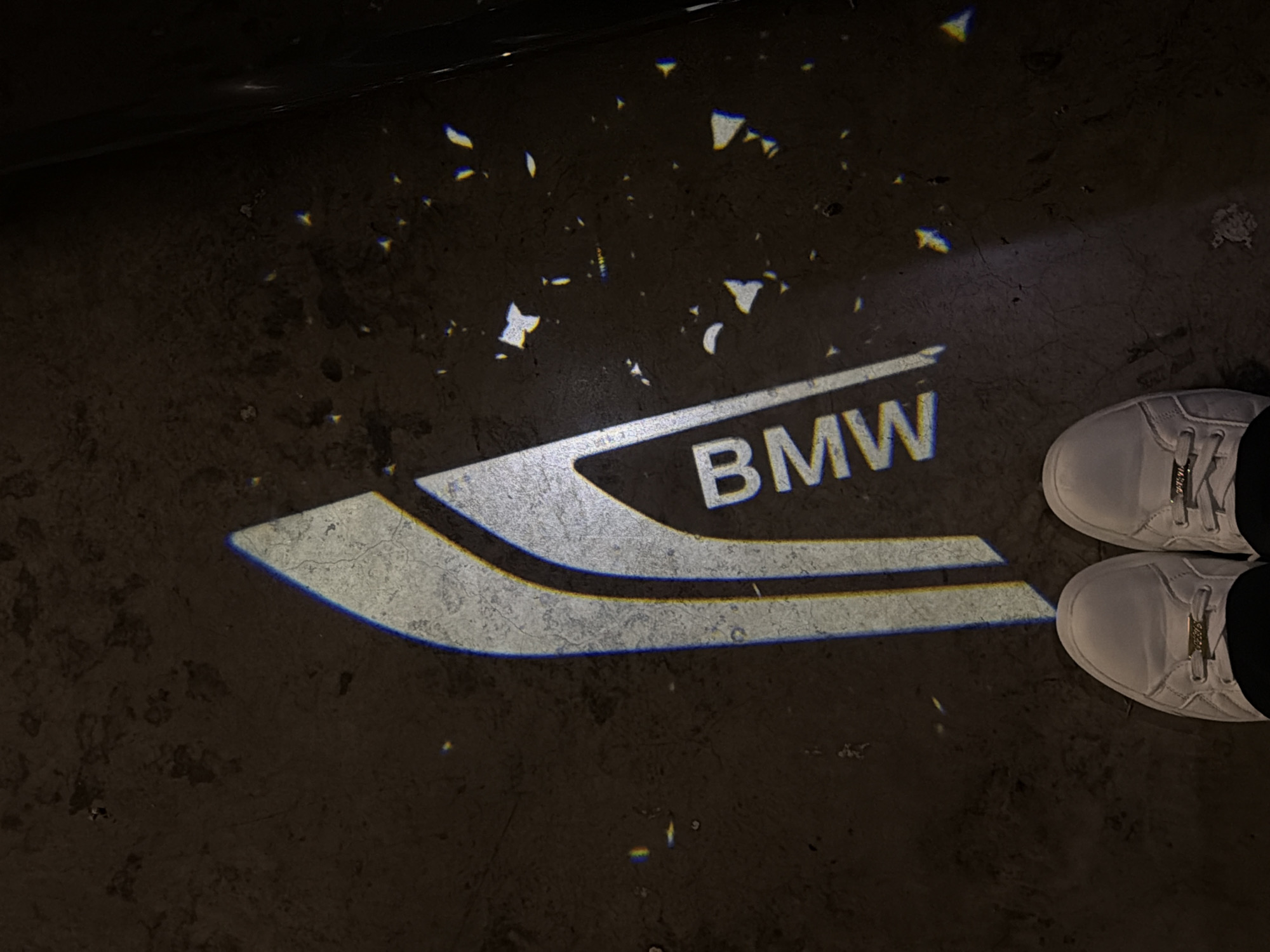 A BMW logo is projected onto a dark, textured concrete floor. The logo is white with blue and yellow outlines, and the letters "BMW" are clearly visible. Small, scattered reflections of light appear on the floor around the logo. In the foreground, the feet of a person wearing white sneakers and dark pants are visible. The scene appears to be indoors, possibly in a garage or showroom.