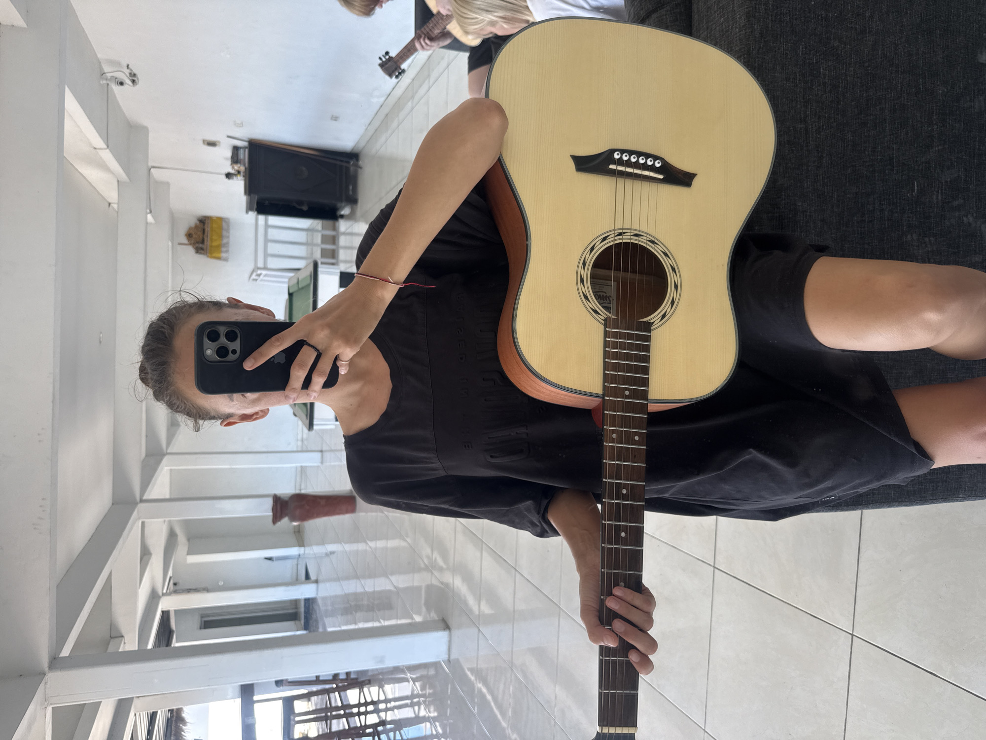A person is holding a guitar in front of a mirror and taking a photo with a smartphone. The person is wearing a black dress and is sitting on a gray couch. The guitar is a light-colored acoustic guitar. The reflection in the mirror shows a white-walled room with a tile floor, columns, and windows. There is also a piece of furniture and another person holding a guitar visible in the reflection. Based on visual elements like the architecture and style, the location is likely in North Kuta, Indonesia.