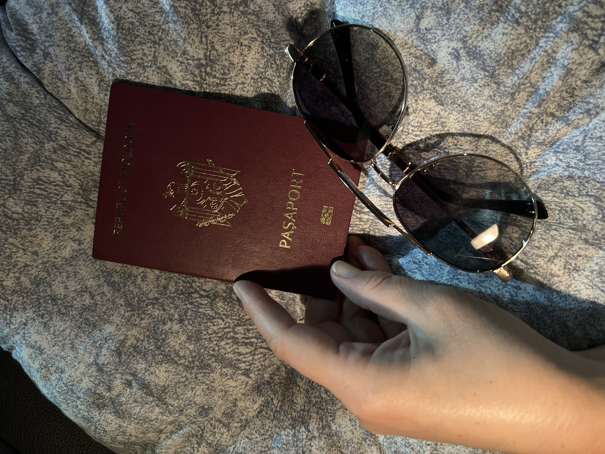 A hand holds a dark red passport with gold lettering and a crest. The passport reads "REPUBLICA MOLDOVA" at the top and "PASAPORT" below the crest. Next to the passport are a pair of gold-rimmed sunglasses with dark lenses. The items are resting on a textured, light blue fabric surface. There is no identifiable location in the city of Giurgiu, Romania.