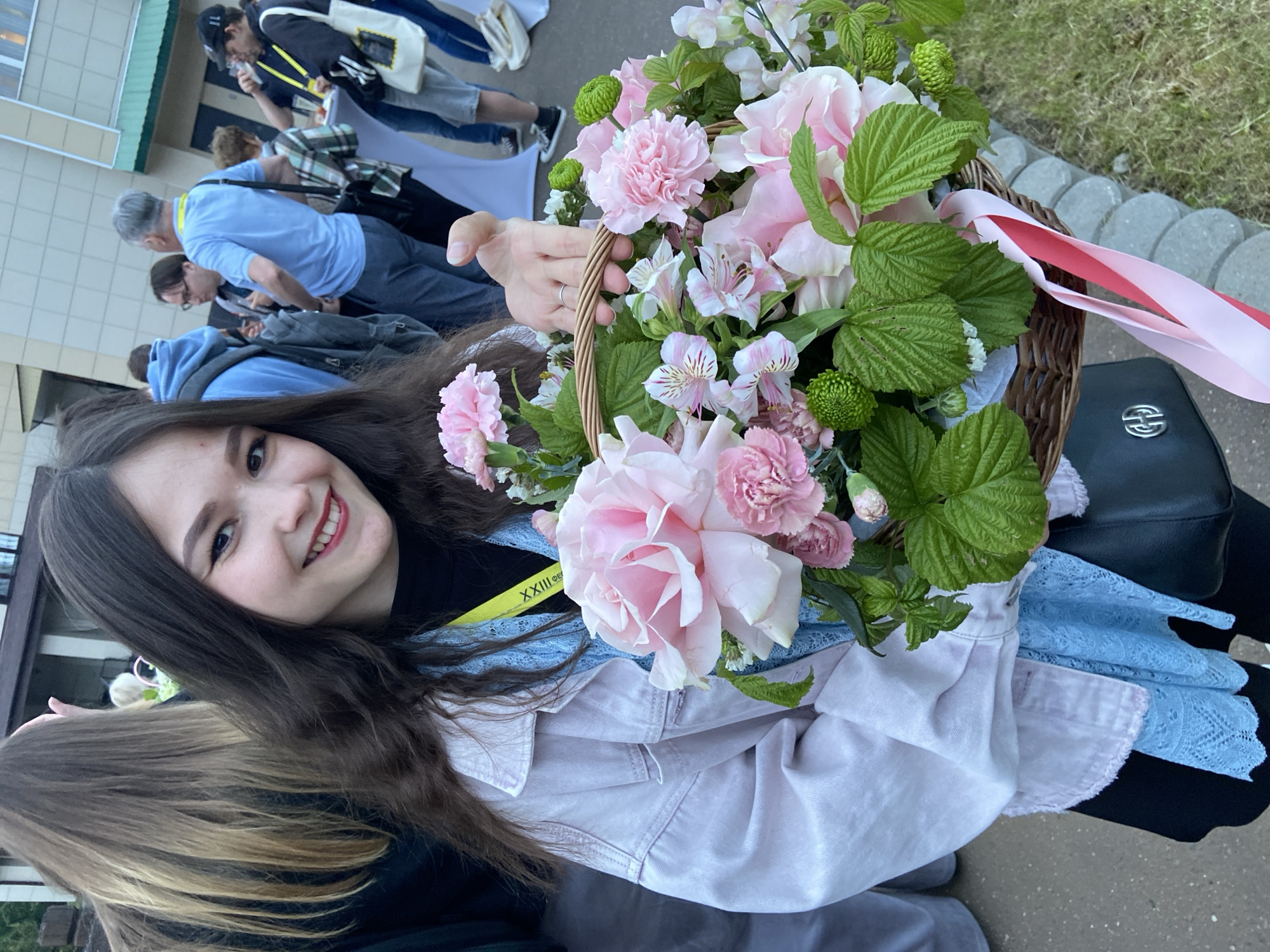 The image, taken in Sergiyev Posad, Russia, features a young woman smiling brightly and holding a decorative flower basket. She has long dark brown hair and is wearing a light-colored (possibly lavender) denim jacket over a blue lace top and a dark underlayer. A yellow lanyard, visible around her neck, shows partial text "XXIII..." and some Cyrillic letters, likely indicating the 23rd iteration of an event or festival.
The woven basket is generously filled with a variety of flowers, primarily soft pink roses, light pink carnations, and white and pink alstroemeria. These are interspersed with vibrant green chrysanthemum-like blooms and lush green leaves, possibly raspberry foliage. Pink and white ribbons are tied to the basket's handle, adding a festive touch.
In the blurred background, several people are gathered, many appearing to wear lanyards, suggesting a public event. A man in a light blue shirt is visible in the mid-ground. The setting includes a light-colored building with a green roofline or awning, situated next to a paved area with grassy sections and grey stone pavers. A black handbag with a silver interlocking logo rests beside the basket. The natural, soft lighting indicates it is daytime, likely under overcast skies, contributing to a cheerful and pleasant atmosphere.