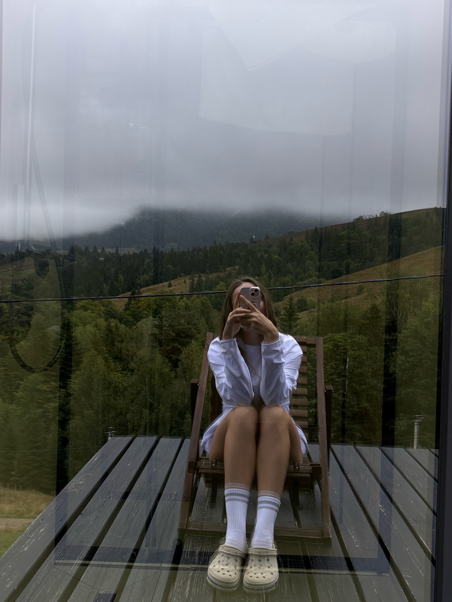 A young woman with long brown hair is sitting in a wooden chair on a wooden deck. She is wearing a white long-sleeved shirt, white socks with gray stripes, and tan crocs. She is holding a pink smartphone in front of her face, as if taking a selfie. The background shows a lush green forest on a hillside under a cloudy sky. The scene appears to be in Krasnyk, Ukraine, a location known for its natural beauty and mountainous terrain. The overall impression is one of a serene and contemplative moment in a natural setting.