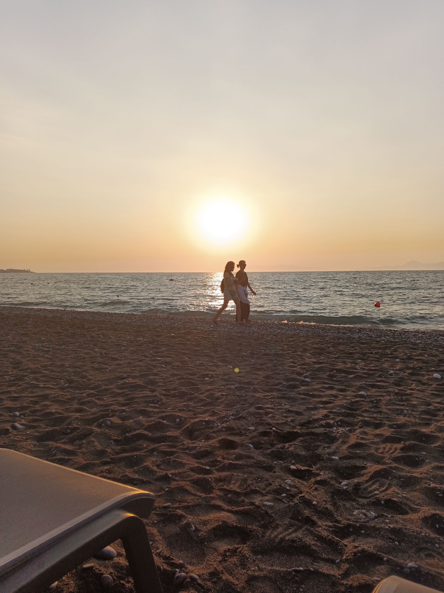 This media file captures a serene beach scene at sunset. The primary subjects are two people silhouetted against the bright, setting sun, walking near the water's edge. One individual appears to be slightly taller and is walking behind the other. The setting is a sandy beach with small pebbles interspersed with the sand. In the distance, the calm ocean stretches out, meeting the horizon where landmasses are faintly visible. The sky is bathed in a warm, golden orange glow, characteristic of a sunset. A single red buoy floats in the water, adding a subtle point of color to the scene. In the foreground, the edge of a lounge chair is visible, hinting at a relaxed atmosphere. The overall mood is peaceful and tranquil, with the soft light and gentle waves contributing to a sense of calm. The time of day is clearly evening, as indicated by the sunset. There is no visible text in the image.