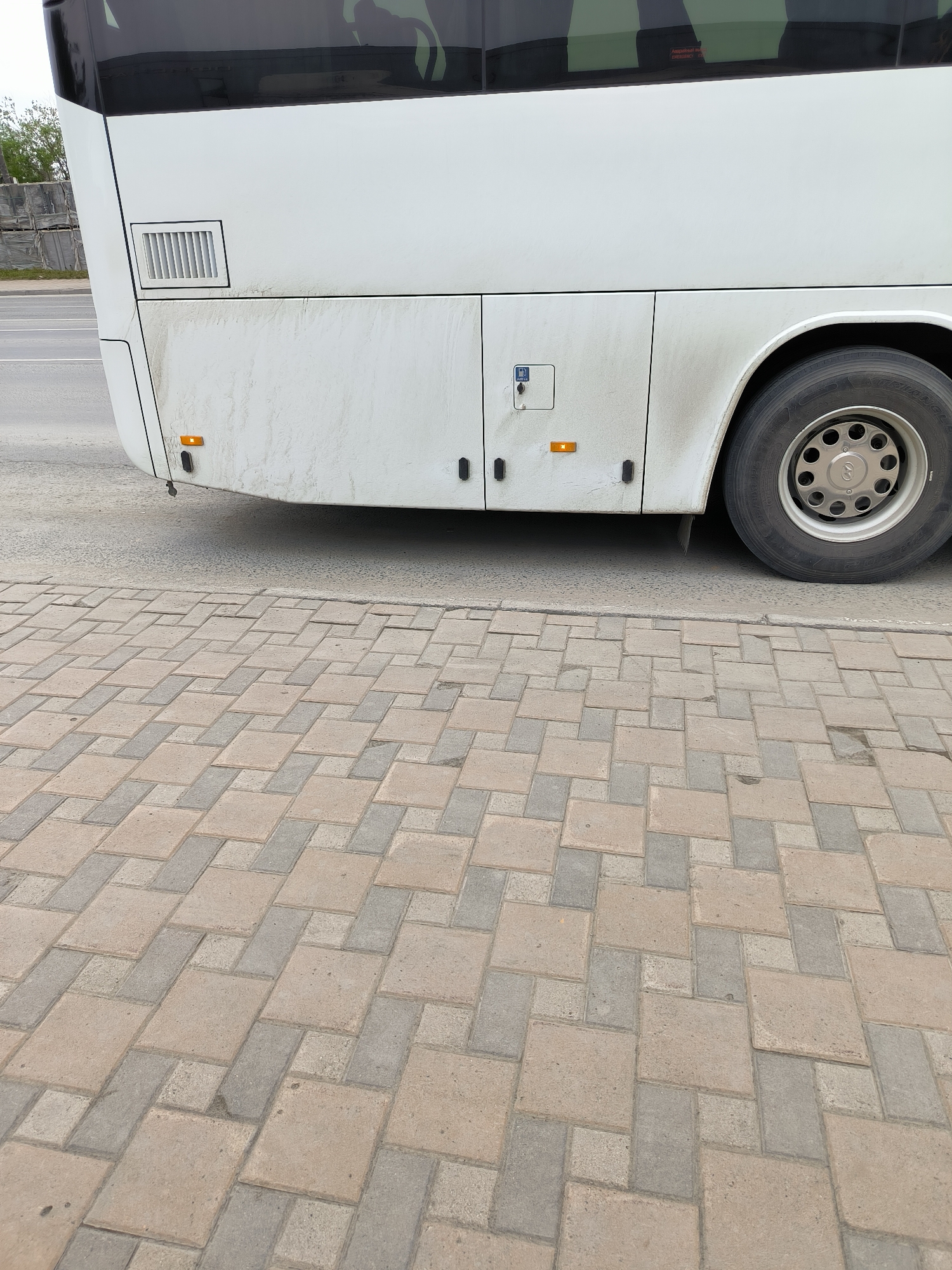 A white bus is parked on a street in Yuzhno-Sakhalinsk, Russia, with a sidewalk paved with beige and gray rectangular bricks in the foreground. The lower portion of the bus is visible, showing the rear wheel, a small orange turn signal, and a fuel cap with a blue symbol. The bus body appears slightly dirty, with streaks of grime running down from the upper sections. The background shows a road and some industrial-looking structures and trees, partially obscured by the bus. The sky is overcast, suggesting a cloudy day.