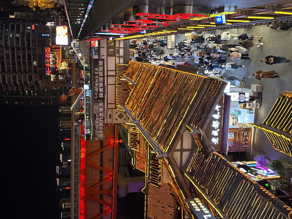 This nighttime image captures a multi-level urban scene in Hongyadong, Yuzhong District, Chongqing, China. The foreground features traditional-style buildings with distinctive tiled roofs, heavily outlined by amber and yellow LED strip lighting, giving them a warm glow. A busy pedestrian street, illuminated by overhead lighting and light spill from adjacent establishments, is visible in the middle ground, populated by a dense crowd of approximately 50-70 individuals. Most pedestrians are walking, observing their surroundings, or congregating in small groups, wearing casual modern attire.
To the immediate right, an elevated walkway or escalator structure, brightly lit with prominent vertical red and yellow neon signs, parallels the pedestrian street. In the mid-distance and background, a massive red truss bridge, identified as the Qiansimen Bridge, spans horizontally across the frame. Its structure is vividly illuminated in red, with streetlights visible on its deck. Above the traditional buildings and below the bridge, an intermediate level features a rooftop area with several people and a large, inflated orange sculpture resembling a figure. Further back, numerous modern high-rise buildings pierce the dark night sky, some displaying illuminated windows and signs. The overall scene is vibrant, dominated by artificial light sources from buildings, signs, and decorative illumination.