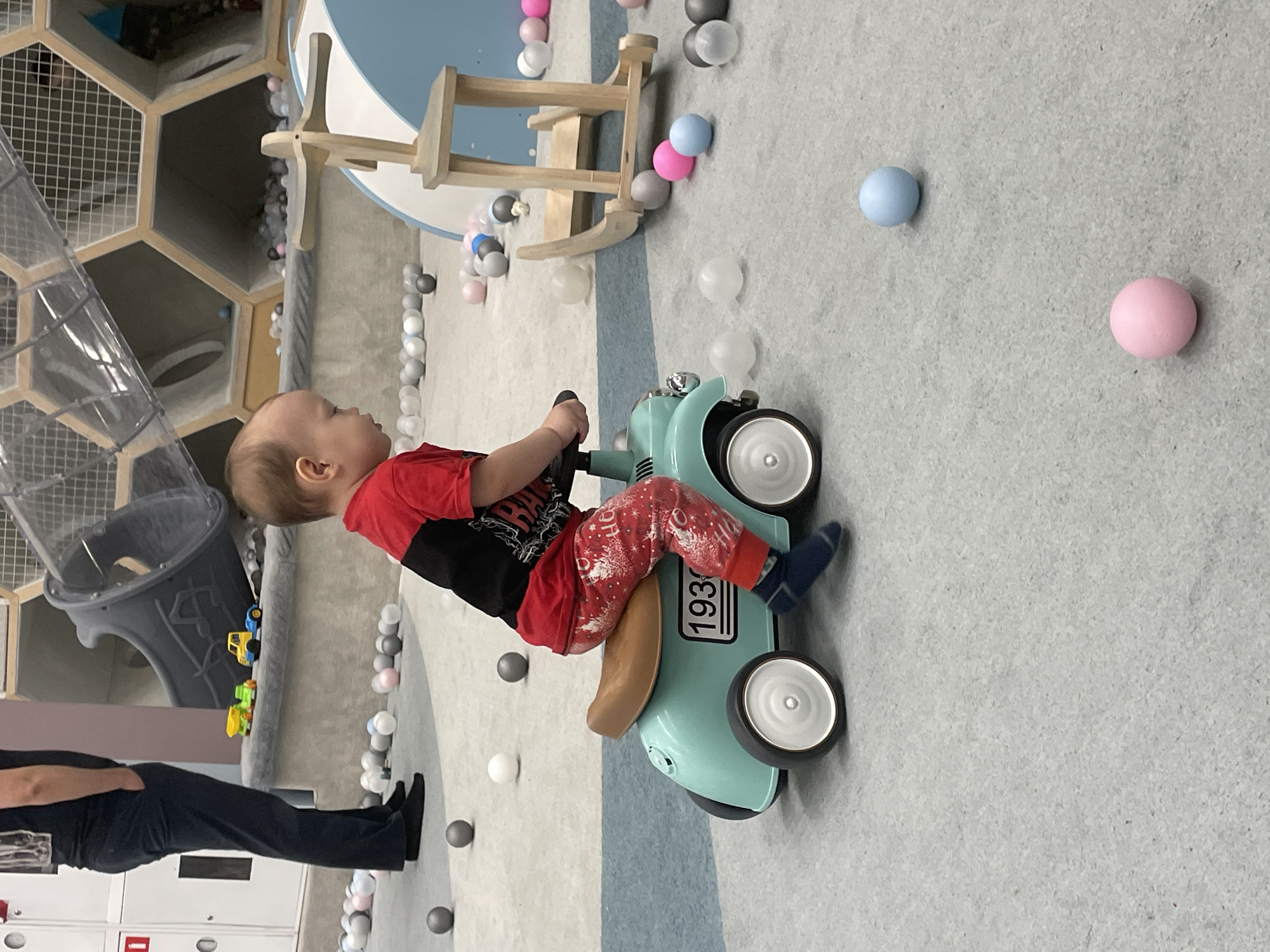 The image shows a baby riding a light blue toy scooter with a brown seat indoors. The baby is wearing a red and black shirt and red and white pants with blue socks. The floor is light gray with blue and gray stripes, and scattered with pink, blue, gray, and white plastic balls. In the background, there is a play structure with a transparent tunnel and hexagonal openings. A person wearing dark clothing is partially visible on the left side of the image. There is a wooden structure near the wall in the background. The location is a children's play area with elements like a ball pit and play structures. The city of Perm, Russia, is not evident from the image.