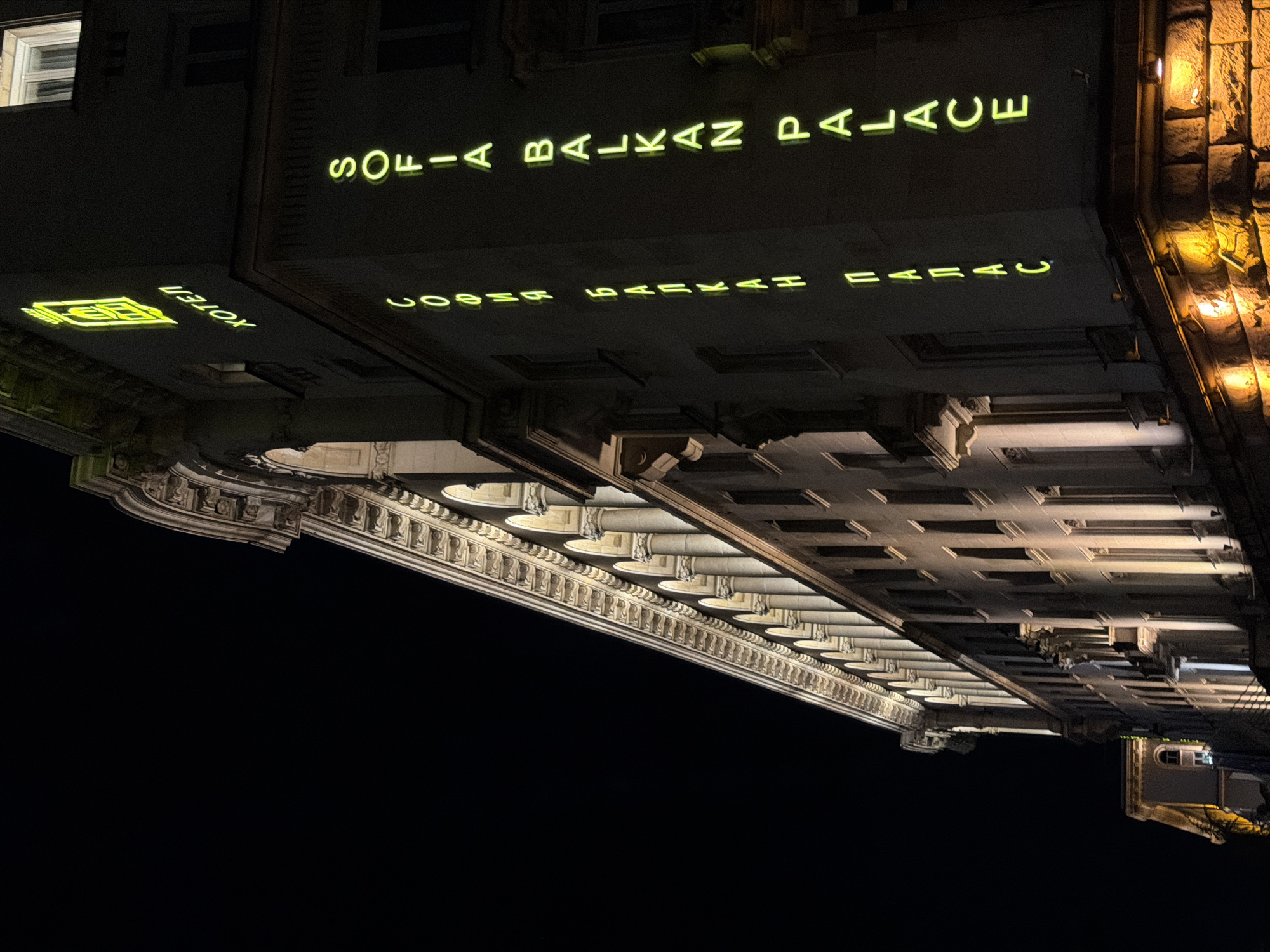 This image captures a grand building at night, viewed from an upward angle. The photograph itself is rotated approximately 90 degrees counter-clockwise.
The subject is a stately, classical-style building, likely a hotel or palace, illuminated by artificial light against a dark night sky. The building features ornate architectural details, including decorative cornices, multiple rows of columns or pilasters along a lower facade, and what appear to be balconies or upper-story windows. The structure is rendered in shades of grey and beige stone, with some sections bathed in warm, golden light from exterior fixtures, highlighting its texture.
Prominently displayed on a darker upper section of the building's facade is a neon sign that reads "SOFIA BALKAN PALACE" in bright yellow-green capital letters. Directly beneath this, a fainter, upside-down reflection of the same text, "SOFIA BALKAN PALACE," is visible, suggesting a reflective surface below the main sign or another sign intended to be viewed from a different angle. Further to the left, another smaller neon sign displays a stylized emblem next to the word "HOTEL," also in the same distinctive yellow-green glow. A solitary illuminated window is visible on the upper left side of the building.
The activity is none; it's a still architectural photograph. The time of day is clearly night, as evidenced by the dark sky and the reliance on artificial lighting to illuminate the building and its signs. The setting is urban, with the grand architecture suggesting a significant historical or luxury establishment. Given the provided context, the location is Sofia, Bulgaria.
