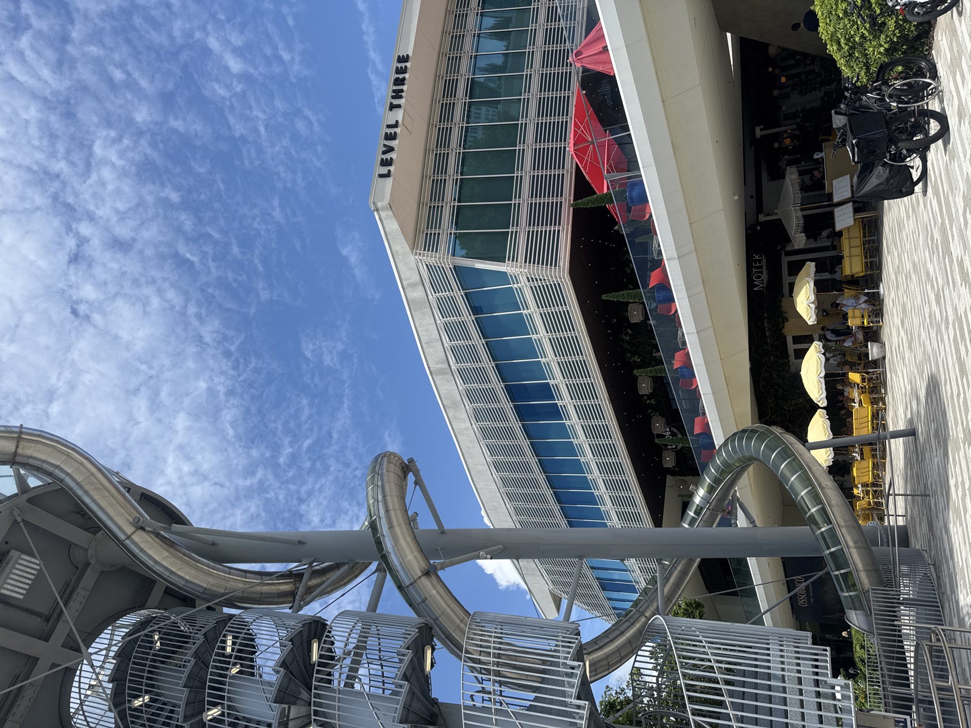This image captures an outdoor view of the Aventura Mall in Aventura, Florida, United States of America. In the foreground, a prominent metal spiral staircase is visible, leading up to a winding, enclosed slide. The slide is constructed from clear material, allowing glimpses of the internal structure. Behind the staircase and slide, a modern, multi-story building dominates the background. This building features a contemporary design with horizontal louvers and large blue-tinted windows. The text "LEVEL THREE" is clearly visible on the top facade of this building.
Below the upper level of the building, a covered walkway or patio area is apparent, furnished with red chairs and large red umbrellas. Further down, an outdoor seating area is set up with yellow tables and chairs, protected by yellow and white striped umbrellas. A sign with the word "MOTEK" is visible above this seating area, indicating a restaurant.
On the ground level, the pavement is laid with a patterned brickwork. Several parked bicycles, including two electric bikes with cargo bags, are situated on the right side of the image, near a lush green hedge. A person is partially visible walking away from the camera in the far right background. The sky is a bright blue with scattered white clouds, suggesting a sunny day. The overall scene depicts a vibrant and contemporary urban public space, likely a shopping or entertainment complex.