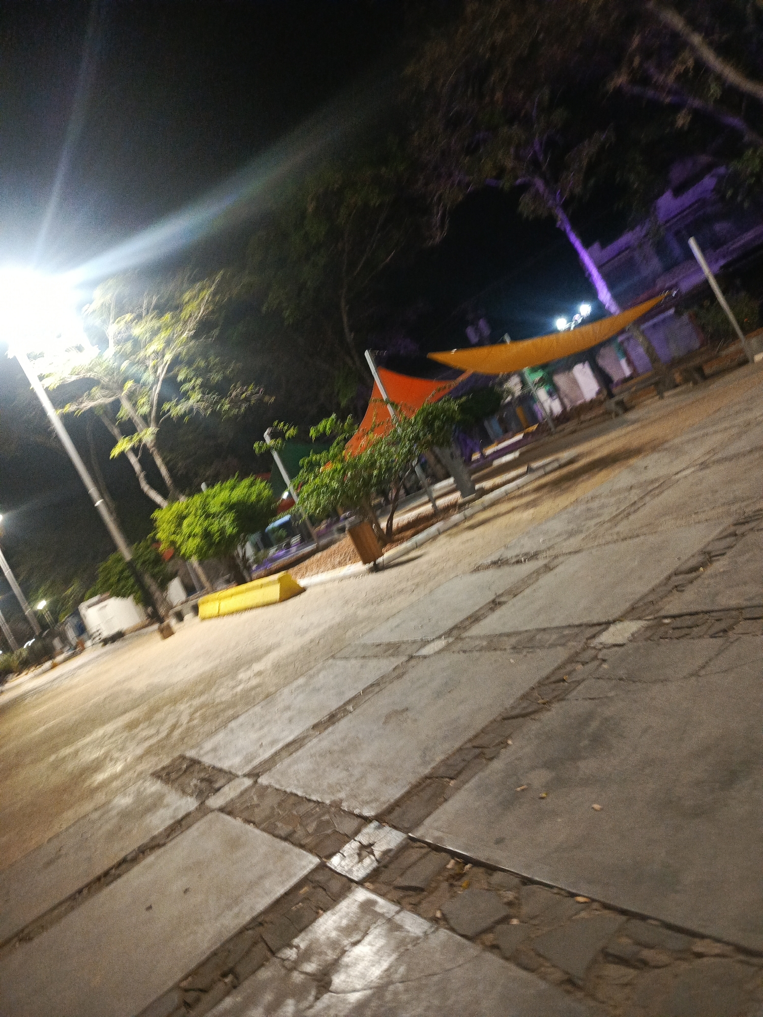 The image depicts an outdoor plaza at night in Juan Griego, Venezuela. A bright light source on the left illuminates trees and the paved ground, which is laid out in a grid pattern with darker inlaid lines. In the background, a bright orange and yellow awning is visible, supported by poles. There are also trees with foliage, some with a purple hue, and structures that appear to be buildings or stalls. A white van is parked to the left, and a yellow rectangular object, possibly a bench or barrier, is situated in the midground. The scene is sparsely populated, with no individuals clearly visible, suggesting a quiet, nighttime ambiance in a public space.