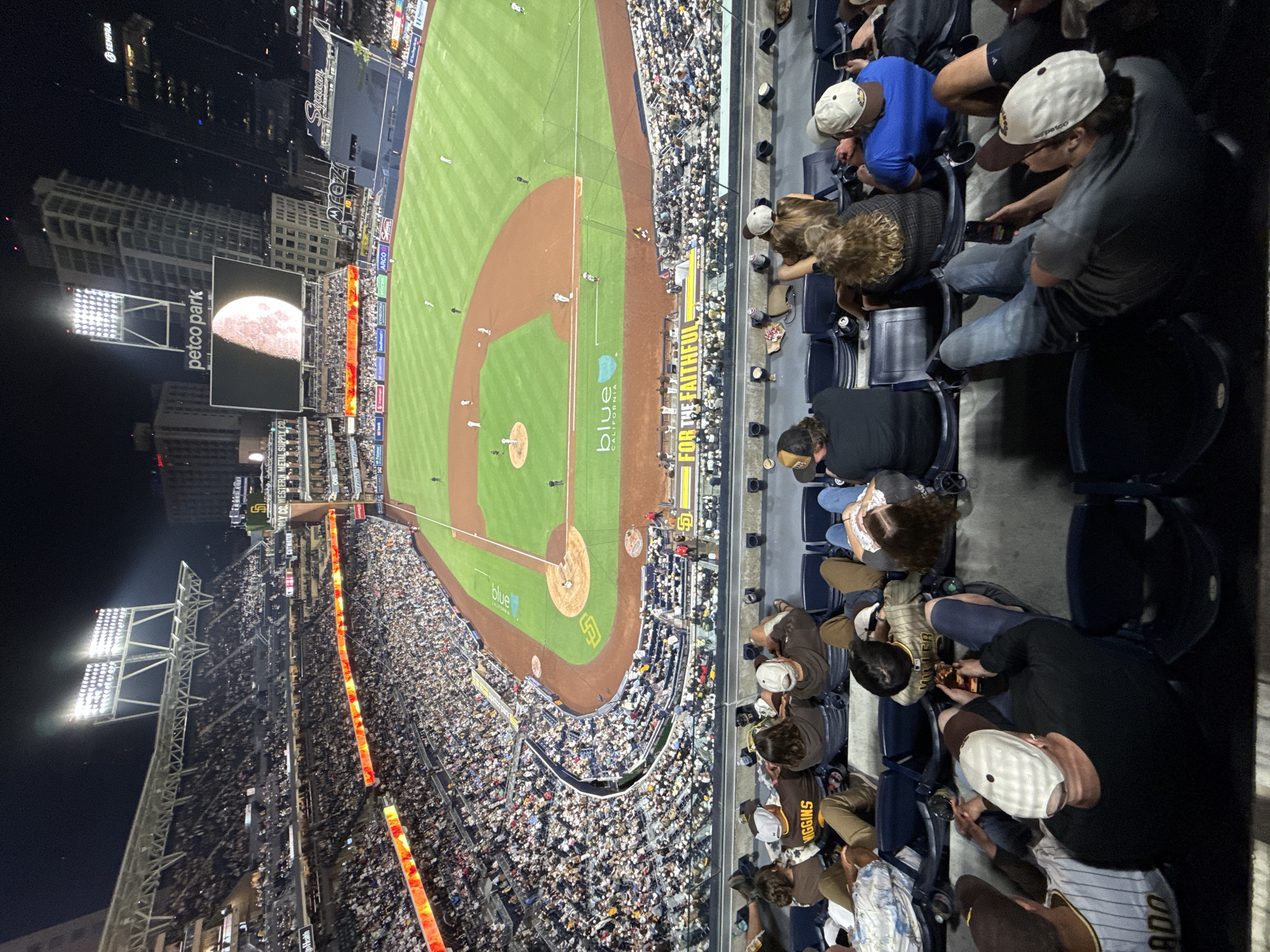 This is a nighttime, high-angle photograph of a baseball game at Petco Park in San Diego, California. The field is illuminated by bright stadium lights, and the stands are filled with a diverse crowd of spectators. The moon is partially visible in the sky, creating a dramatic backdrop.
The image captures the essence of a baseball game atmosphere, with fans engaged in watching the sport. Many individuals are seated, some holding phones, likely capturing memories or checking scores. The overall mood appears to be one of enjoyment and shared experience.
Visible text includes "petco park," "blue CALIFORNIA," "FOR THE FAITHFUL," and several corporate sponsorships such as "ARCO" and "SEMPRA." The scoreboard displays game information and advertisements. The urban skyline of San Diego is visible in the background, adding to the setting's context. The photograph is characterized by its vibrant colors, sharp details, and the dynamic interplay of light and shadow.