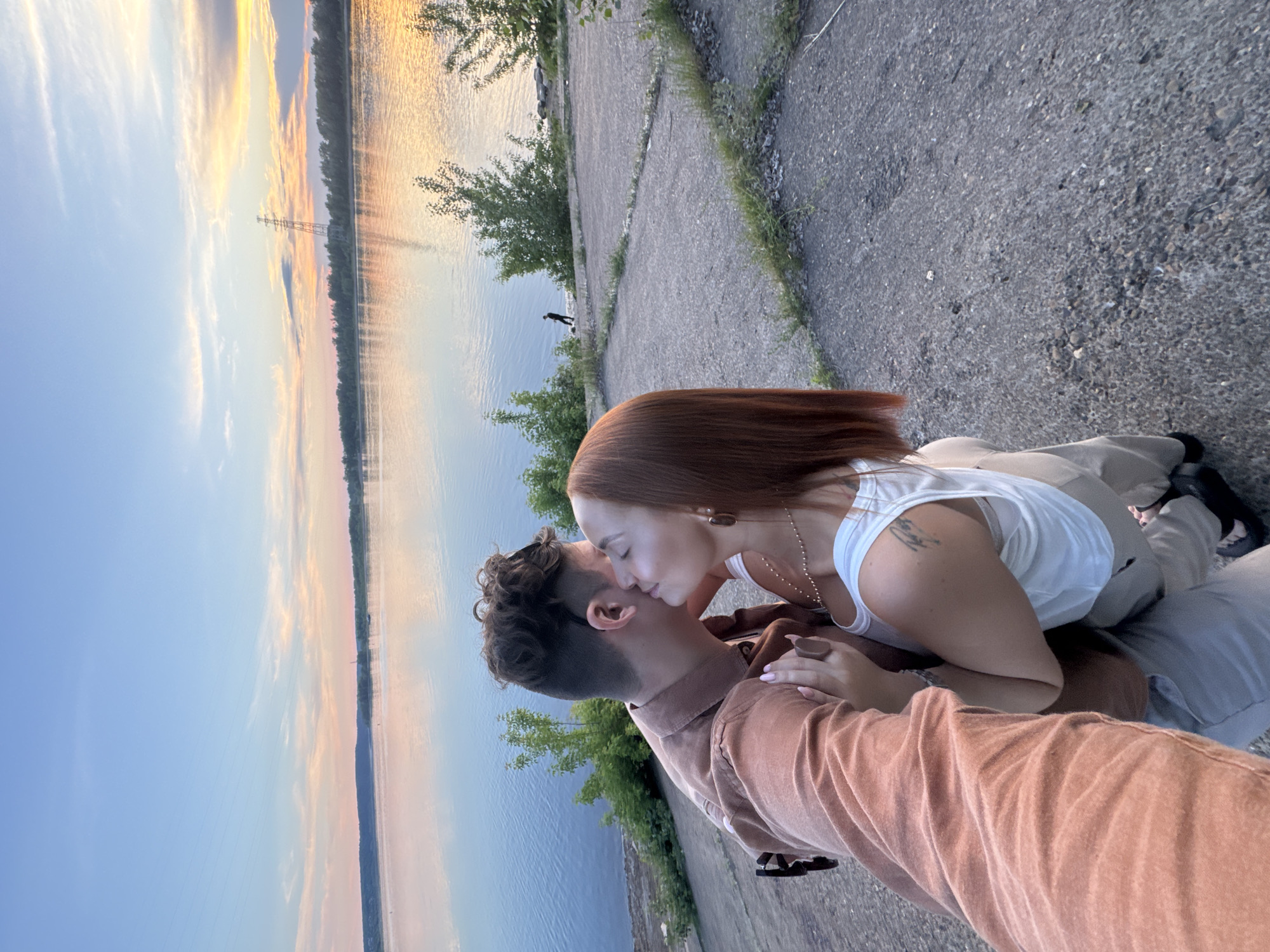 A couple is kissing on a paved surface next to a body of water at sunset. The man is wearing a brown button-up shirt and the woman is wearing a white tank top. The water is calm and reflects the colors of the sunset, with a line of trees visible on the opposite shore. In the distance, a power line tower is visible against the colorful sky. There is a single person standing by the water's edge in the background. The paved area appears to be an outdoor gathering space or overlook.