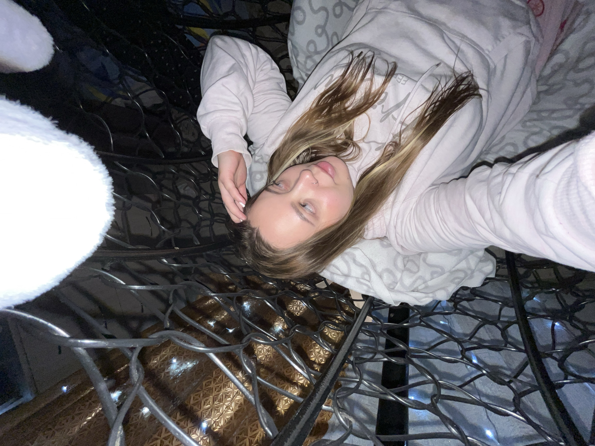A young woman with long, blonde hair is lying down on a gray patterned blanket. She is wearing a light pink hoodie. The perspective is an overhead shot looking down through a metal grid-like structure. The background behind the grid is blurred and shows hints of patterned paper. The photo was taken indoors. There is no confirmed evidence to indicate it was taken in the city of Novopokrovskaya, Russia.