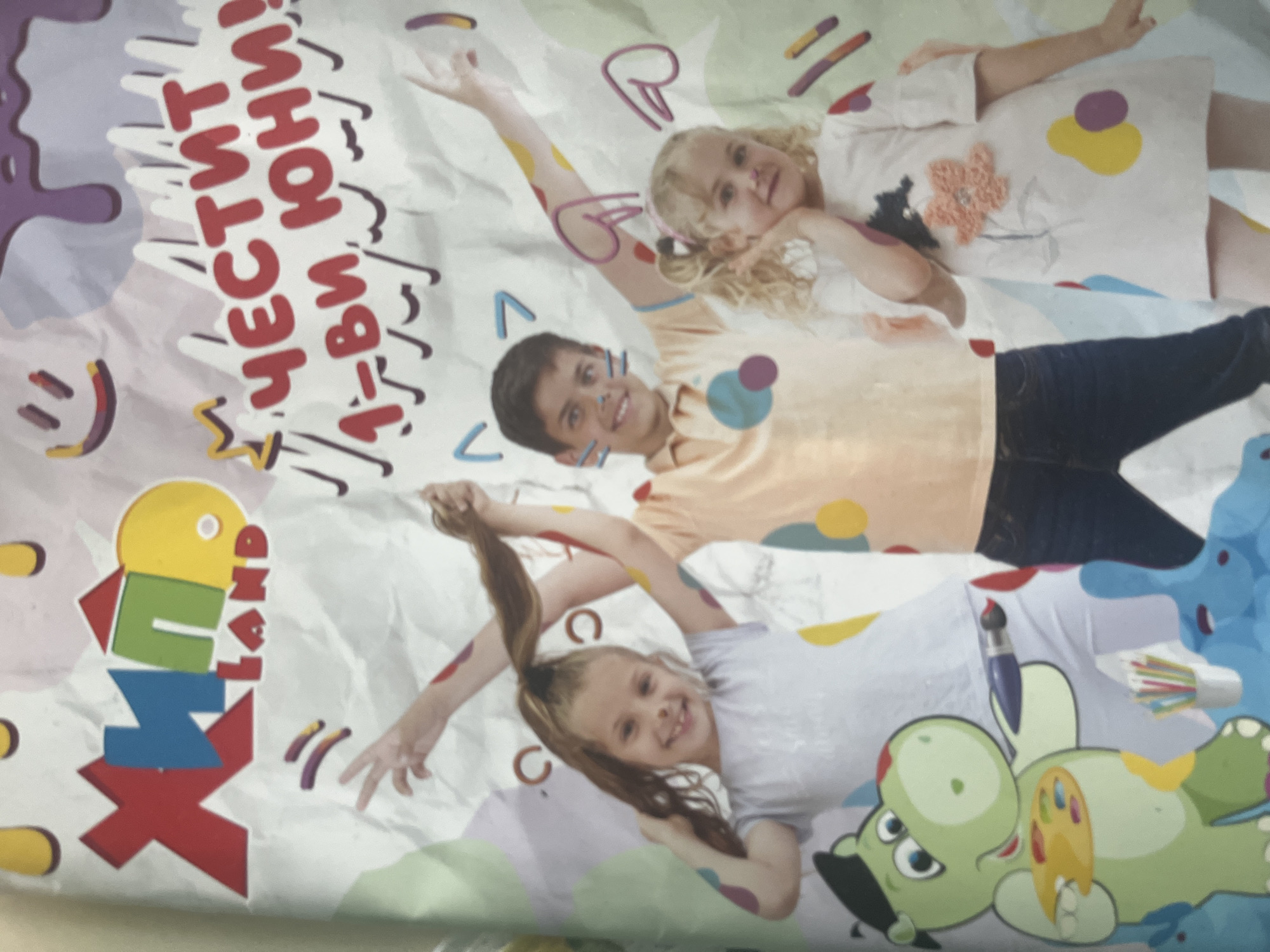 The image depicts a printed advertisement for a "Land" themed play area with the words "ЧЕСТИТ 1-ви ЮНИ!" (Happy 1st of June!) in red. The advertisement features three children lying down and appearing to paint on a background that appears to be crumpled paper. A girl with blonde curly hair is resting her head on the shoulder of a boy, who is smiling and has his arm raised. Below them, a girl with brown hair is smiling and also has her arm raised. A cartoon dinosaur-like creature holding a palette of colors is present as well. This is the image found in Pokrovnik, Bulgaria.