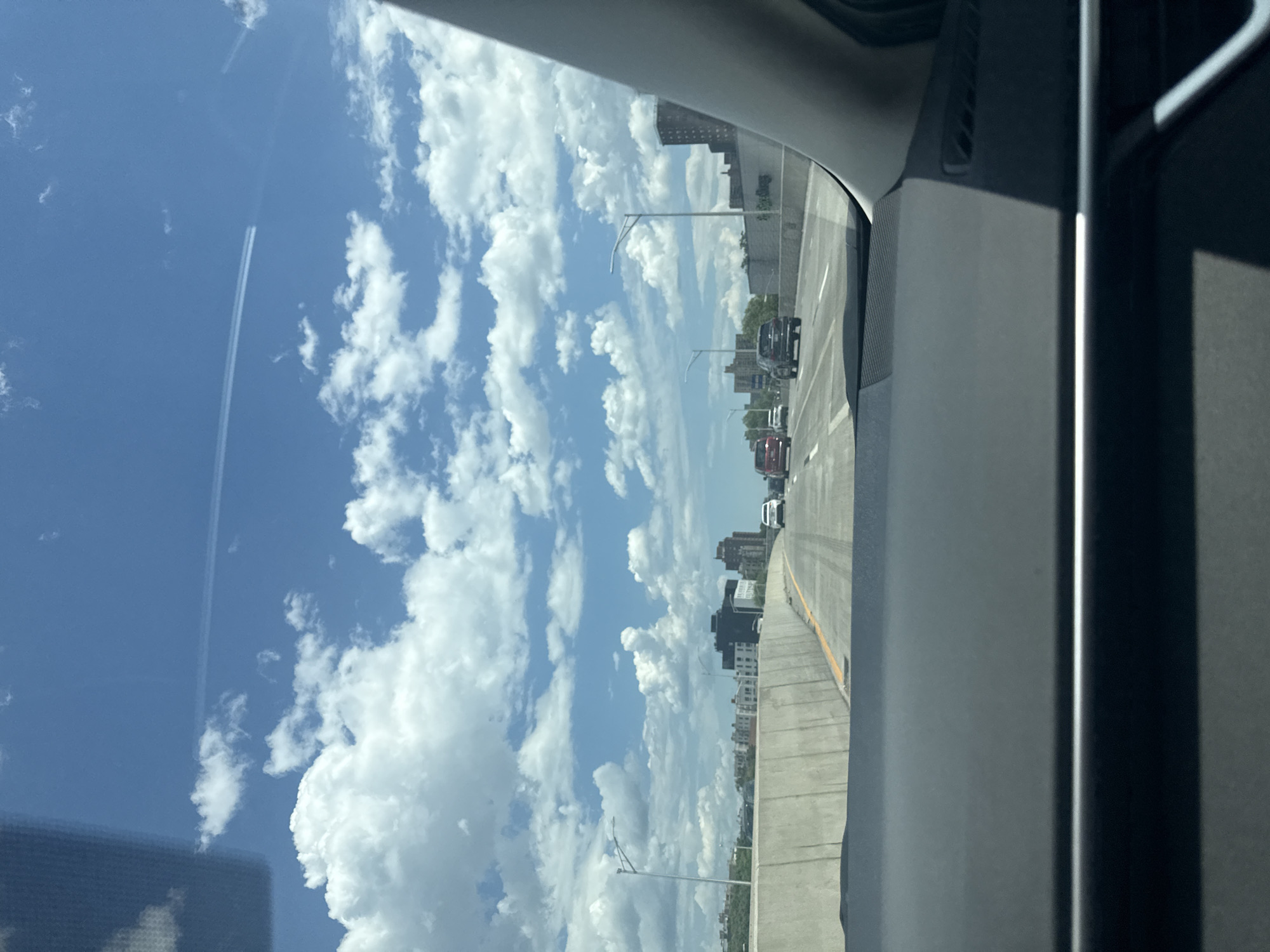 The image provides a forward-facing perspective from the interior of a vehicle on a multi-lane highway, likely elevated due to the prominent concrete barrier. The sky is a vibrant blue, filled with numerous scattered white cumulus clouds. A distinct, linear white contrail from an aircraft is visible high in the upper sky, extending diagonally from the upper left towards the upper right.
On the left side of the highway, a tall, grey concrete barrier runs parallel to the road, with vertical panel lines visible on its surface. Beyond this barrier, a dense urban landscape unfolds, featuring various multi-story buildings, some appearing residential with brick facades, others taller and darker, suggesting commercial or mixed-use structures. Modern L-shaped streetlights with horizontal fixtures are positioned along the highway, particularly visible on the left.
The highway ahead shows moderate traffic. In the immediate lanes, a dark-colored SUV is visible, followed further down by a white sedan in the right lane and a red SUV in the left lane. Additional vehicles are discernible further in the distance.
The lower portion of the image is dominated by the dark grey dashboard of the vehicle from which the photograph was taken. A horizontal chrome or silver trim strip runs across the dashboard. The right A-pillar of the vehicle is visible along the right edge of the frame, partially obscuring the view.
While the urban characteristics, architectural styles, and highway infrastructure are consistent with an urban highway setting within New York City, specific identifying landmarks that would allow for a precise pinpointing of the location within the city are not discernible in this image.