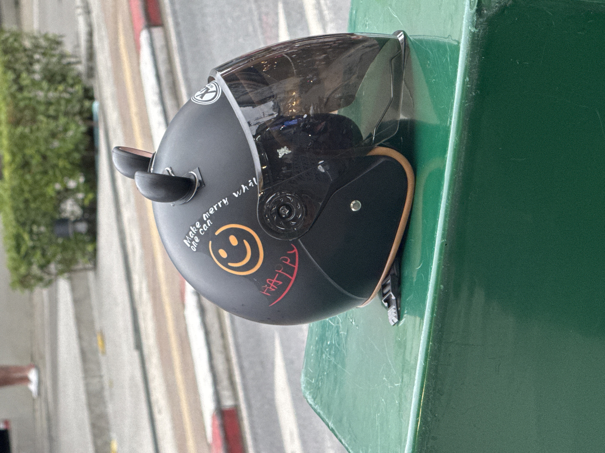 A black motorcycle helmet with two small, black, ear-like accessories is positioned on a green, metallic surface. The helmet features a smoked visor and is decorated with a smiley face, the text "Make merry, one can," and the word "HAPPY" written in red. A person's legs, wearing white shoes, are visible in the upper left background, suggesting they are standing on a tiered walkway or steps. The environment appears to be outdoors, with a street and some green foliage in the background. The scene is set in Karon, Thailand.