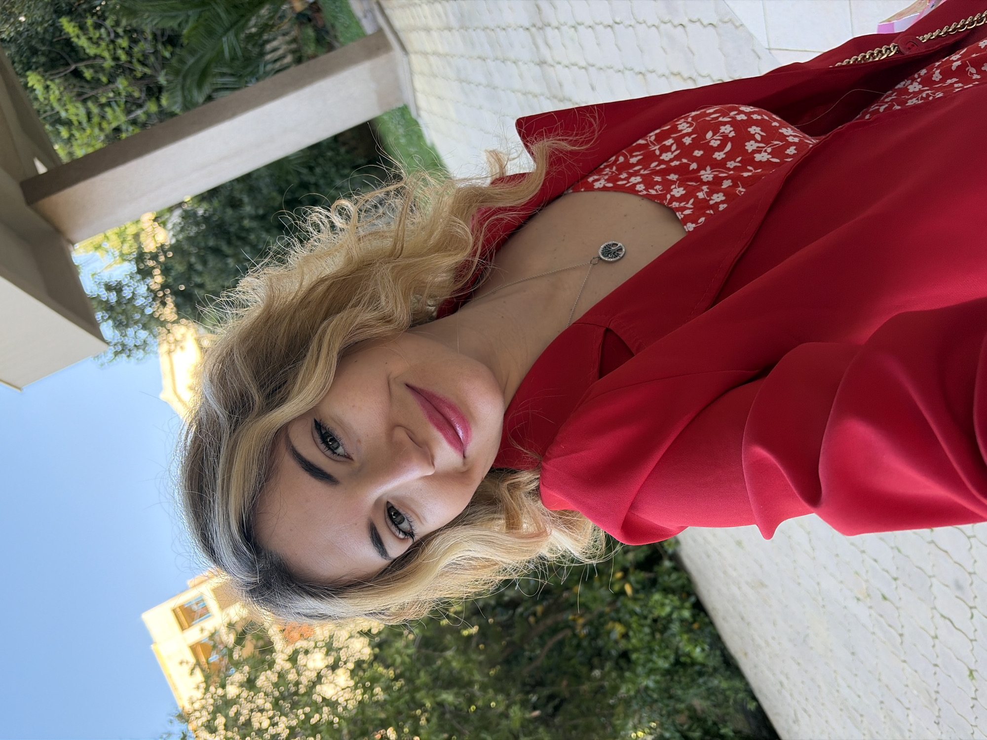 A woman is posing for a picture outdoors. She is wearing a red jacket over a red floral dress. She is also wearing a silver necklace. The location appears to be in Budva, Montenegro, with stone pavement visible and greenery. Buildings are partially visible in the background under a clear blue sky.