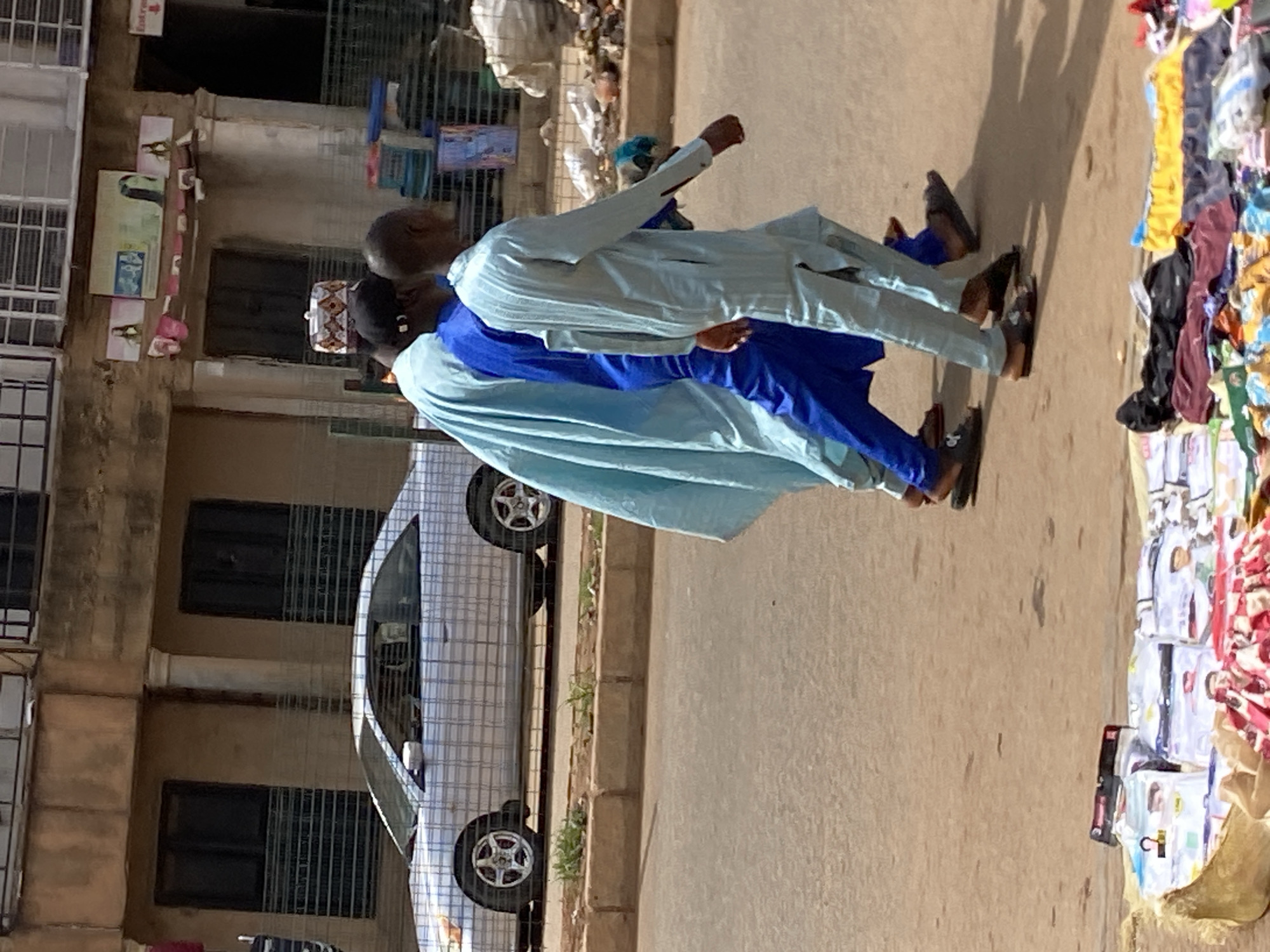 The image shows two men walking along a sidewalk in Bukuru, Nigeria. The man in front is wearing a light blue outfit while the man behind him is wearing a dark blue outfit. They are walking past a car parked alongside a building. Beside them, there is a display of items for sale on the sidewalk.