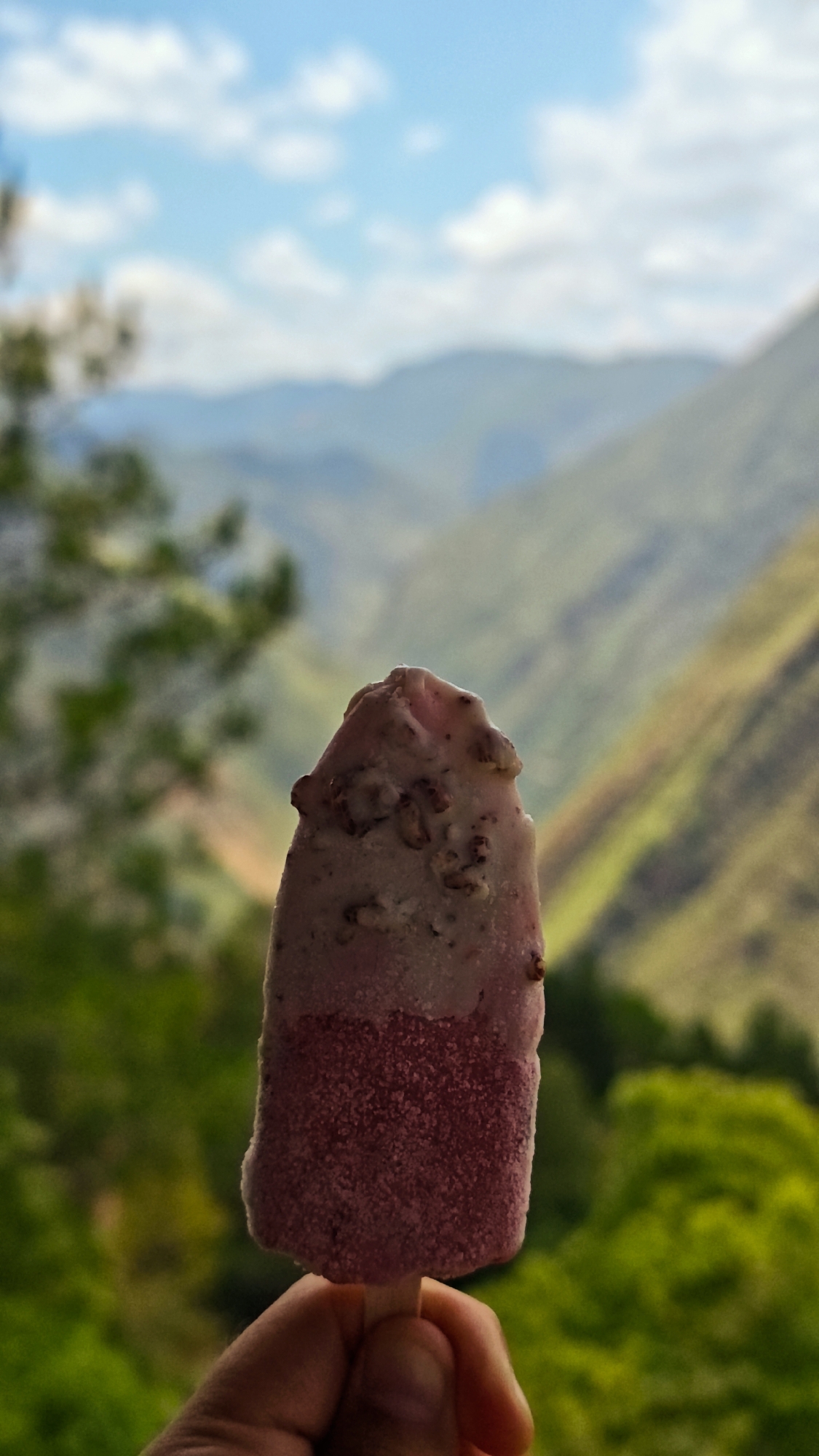 A hand holds a three-layered popsicle against a blurred mountain landscape. The popsicle has a dark pink bottom, a lighter pink middle, and a mottled white and pink top with dark bits. The background shows green trees and a hazy mountain range under a blue sky with scattered clouds. The location is inferred from the prompt to be Lũng Cú Commune, Vietnam.