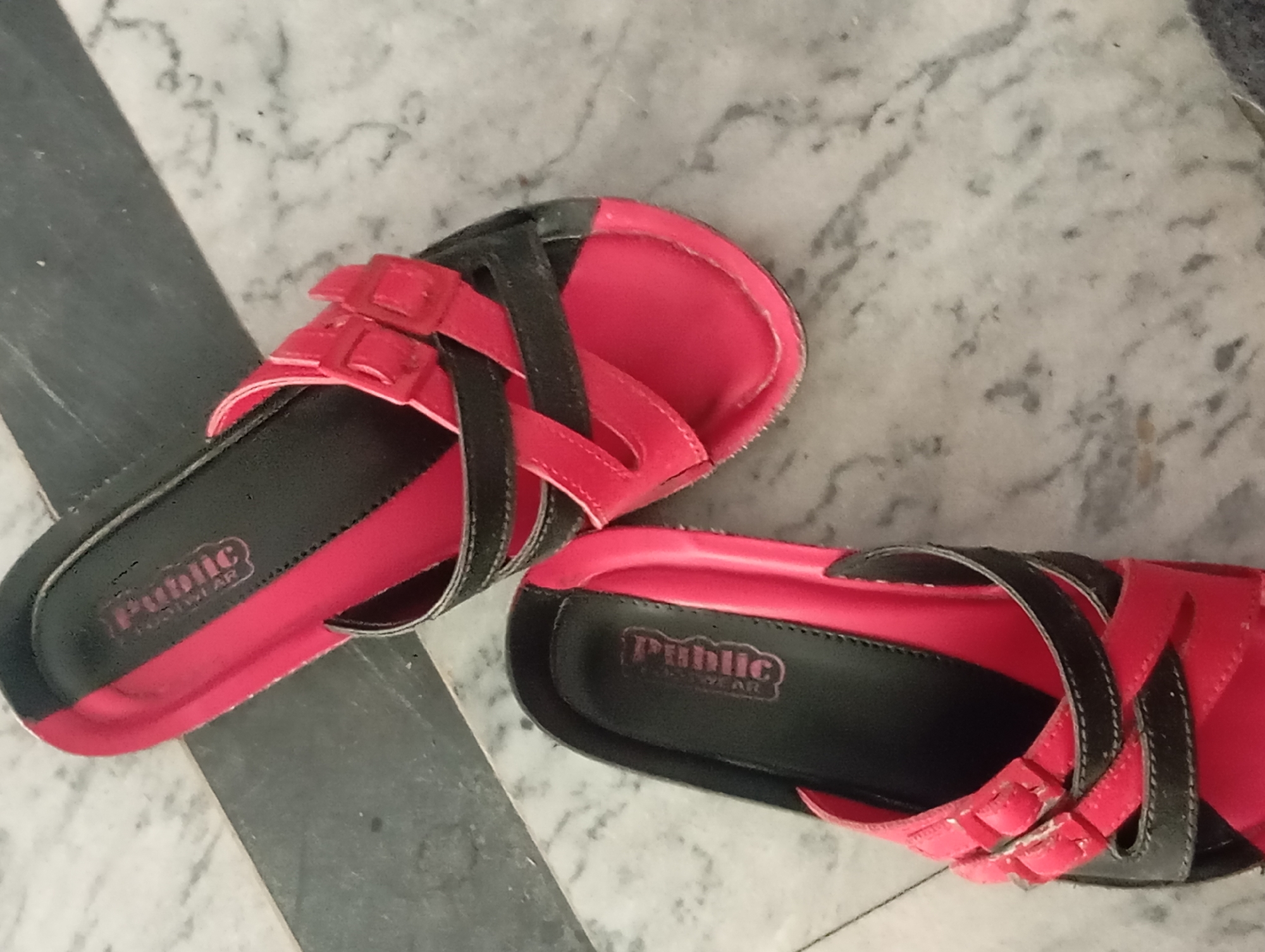 Two pink and black women's sandals are lying on a marble floor. The sandals have crisscrossed straps and buckles. The brand name "Public Footwear" is visible on the insole of both sandals. One sandal is on the left side of the image and the other is on the right.