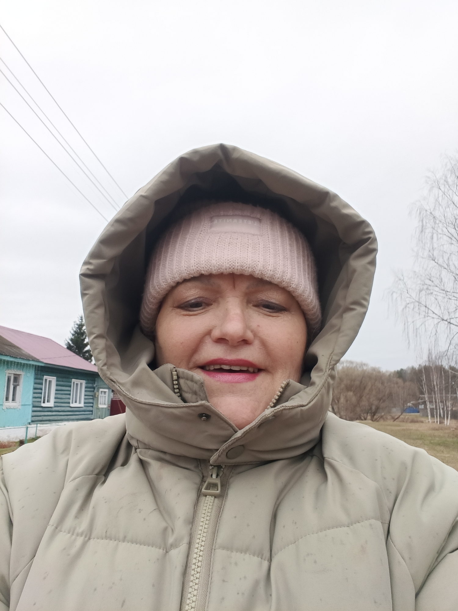 A selfie-style photograph shows a middle-aged woman wearing a light-colored, puffy winter coat with the hood up. Underneath the hood, she is wearing a pink knit hat. The woman has a fair complexion and is wearing lipstick. The background reveals a rural scene. To the left, a blue and white wooden house with a red roof is visible. In the distance, bare trees stand against a cloudy sky. Power lines can be seen in the upper-left corner of the image. The photograph appears to have been taken outdoors. The location is Красный Маяк, Russia.