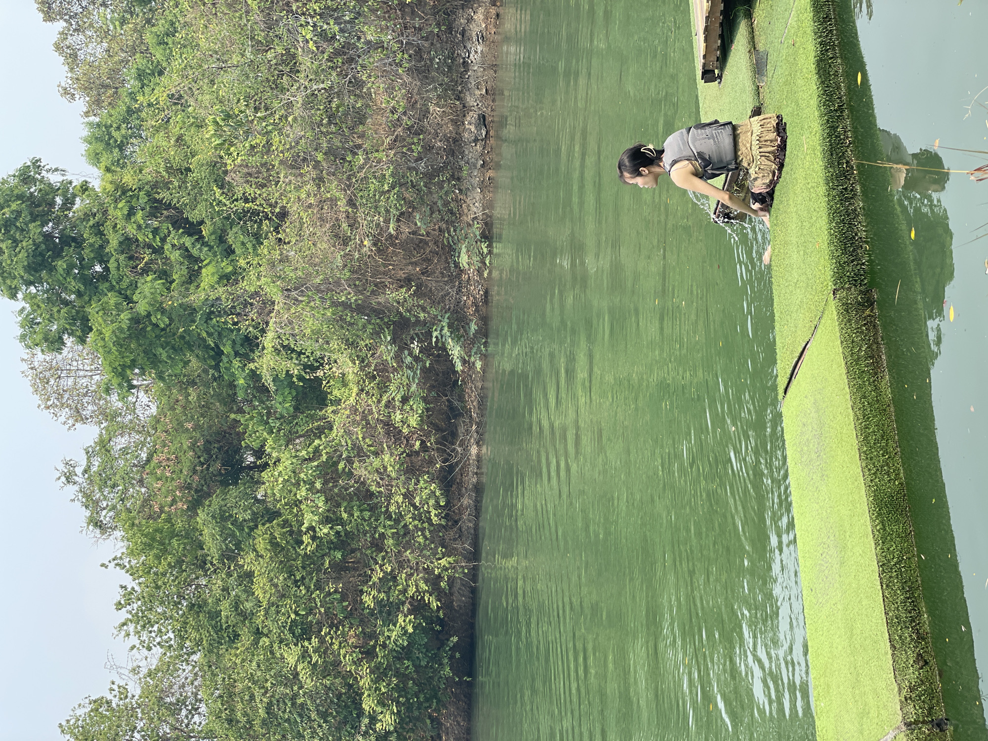 A young woman in a grey top and a camouflage life vest is standing on a platform covered in artificial turf, leaning over and reaching into a body of green water. The platform appears to be a floating dock or pier. The water reflects the sky and vegetation. On the opposite side of the water from the woman, a bank is densely covered with green foliage and trees. The image was taken outdoors, presumably in the location Tambon Nam Phrae, Thailand.