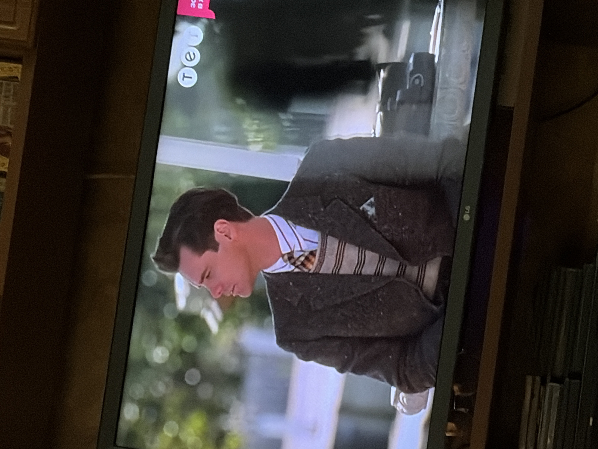 The image shows a television screen displaying a scene with a man wearing a suit and tie, with a distinctive hairstyle. The man is looking to his left. On the screen, in the upper right corner, there is a logo that appears to be "tet". The television screen is placed on a wooden shelf, which is part of a furniture unit. Below the television, there are stacks of books or magazines. The background on the screen suggests an outdoor setting with blurred greenery and a white structure. The overall impression is a still from a movie or TV show. The location in the city of Osolynka, Ukraine, is not discernible from the image itself.