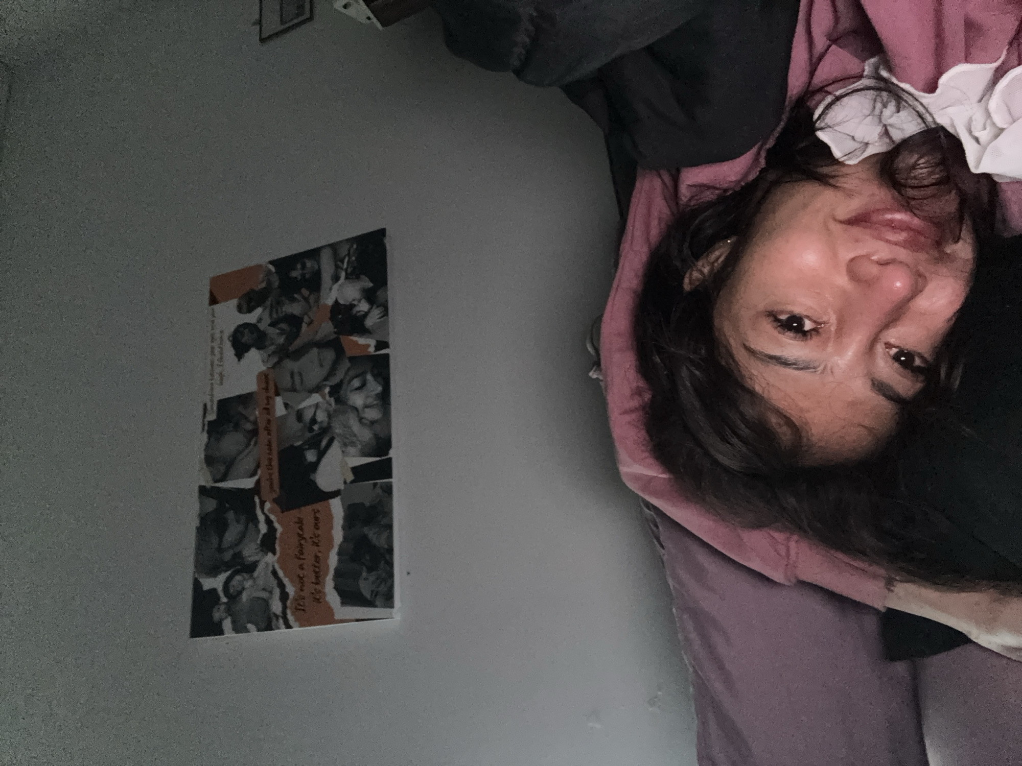 The image shows a person lying in a bed, with their head at the top right of the frame. The person has dark hair and appears to be wearing a pink top with a white collar. To the left of the person is a wall, on which a collage of black and white photographs is hung. The collage has some text on it, including the phrases "Jore the cabe after at hay chase" and "It's not a fairytale, it's better, it's ours." The overall environment appears to be a bedroom. The location and city are unknown.