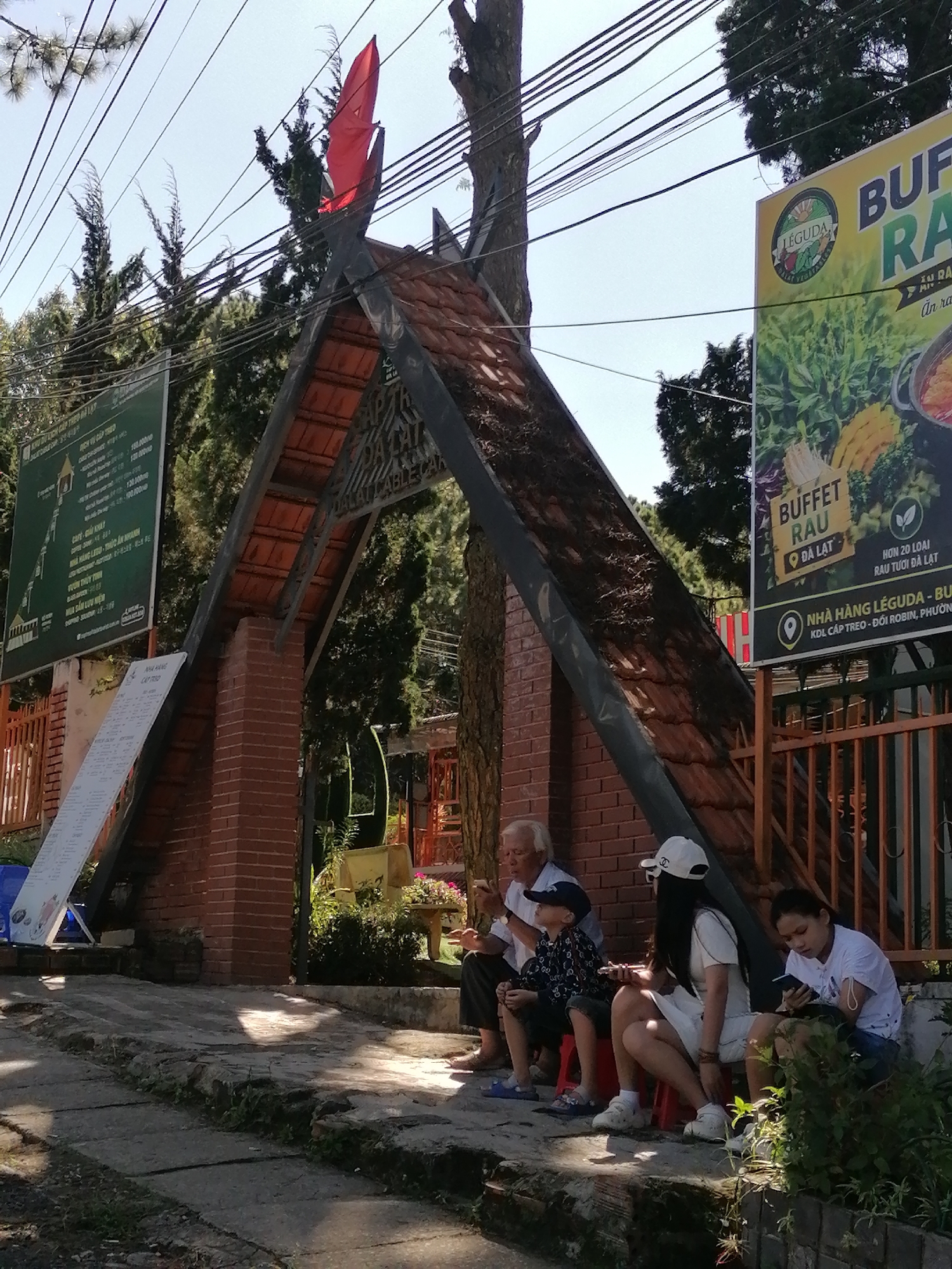 In Da Lat, Vietnam, a family of four is seated on red stools in front of a large A-frame structure with a red tiled roof. The structure appears to be an entrance or a display archway adorned with wood carvings and a red flag at its peak. To the left of the archway, a green sign lists prices for various attractions. To the right, a yellow and green billboard advertises "BUFFET RAU DA LAT." The family consists of an elderly man, a young boy wearing a blue baseball cap and patterned shirt, a woman in a white cap and dress, and a young woman in a white t-shirt. All are looking at their phones or engaged in other activities. The environment is sunny with dappled shade cast by overhead trees and power lines. The ground is paved with stone and concrete. The overall scene suggests a tourist location.