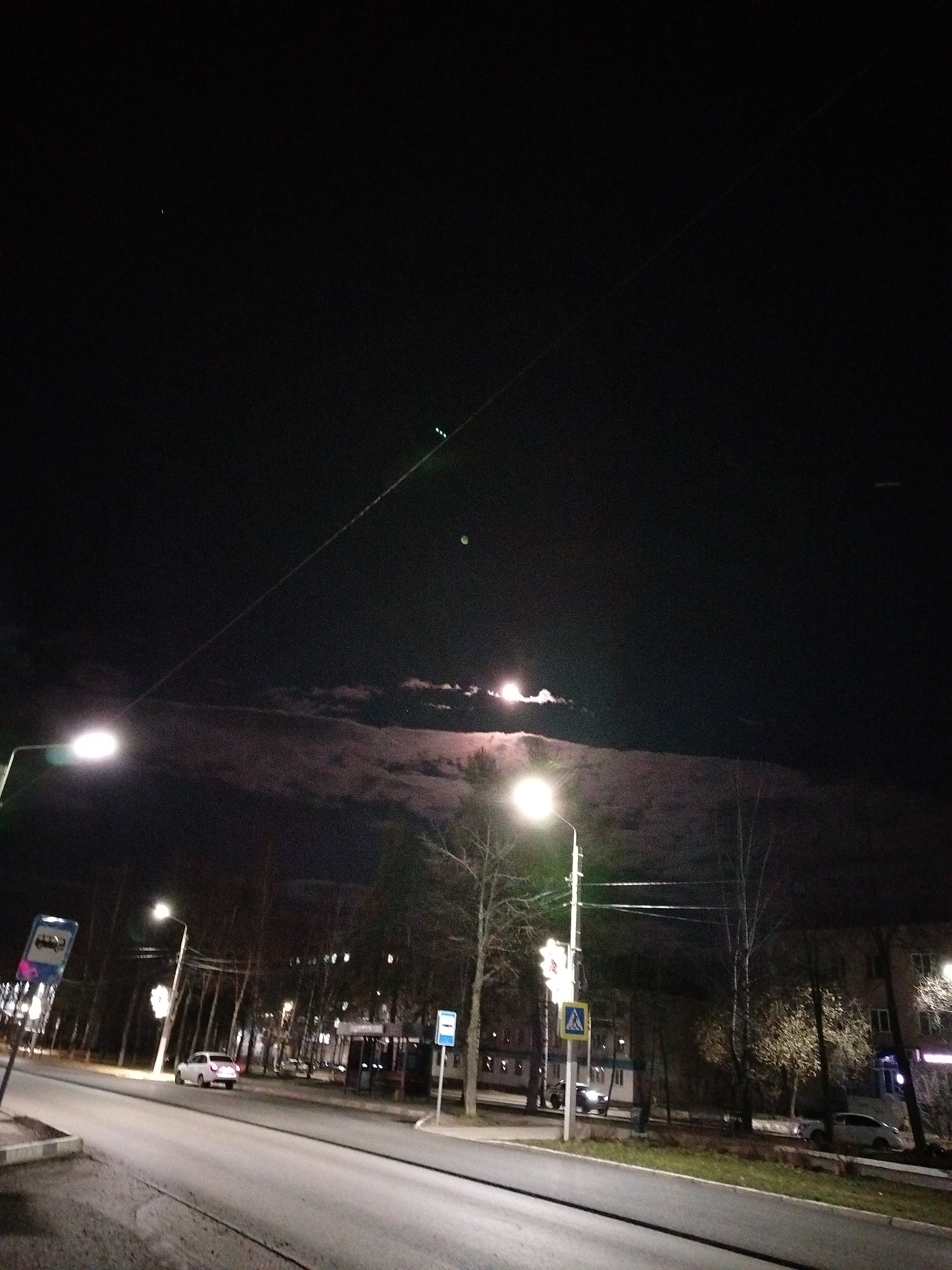 The image captures a nocturnal street scene in Chaikovsky, Russia. The sky, occupying the upper half of the frame, is dark, with a luminous celestial body, likely the moon, visible in the upper right quadrant, partially obscured by wispy clouds that glow brightly from an unseen light source. Several long, thin lines, possibly utility cables or wires, stretch across the sky, with small green lights attached to one line in the upper right.
The street, appearing wet or reflective, extends into the distance, bordered by sidewalks and bare deciduous trees. Multiple streetlights illuminate the road and surroundings with bright, warm glows. A prominent streetlight on the left casts a wide beam, while another bright streetlight is visible further down the street on the right.
Along the road, a white car is parked or moving slowly towards the right, and other vehicles can be seen further in the background. Buildings with illuminated windows line the street, primarily on the right side, suggesting a residential or commercial area.
Visible road signage includes two blue rectangular bus stop signs with white bus icons – one prominently on the left and another further down the street on the right. Near the right-hand streetlight, a yellow diamond-shaped sign with a black pedestrian crossing symbol indicates a crosswalk. The overall atmosphere is quiet and still, typical of a late-night urban environment. No real-world text from within the scene is clearly legible.