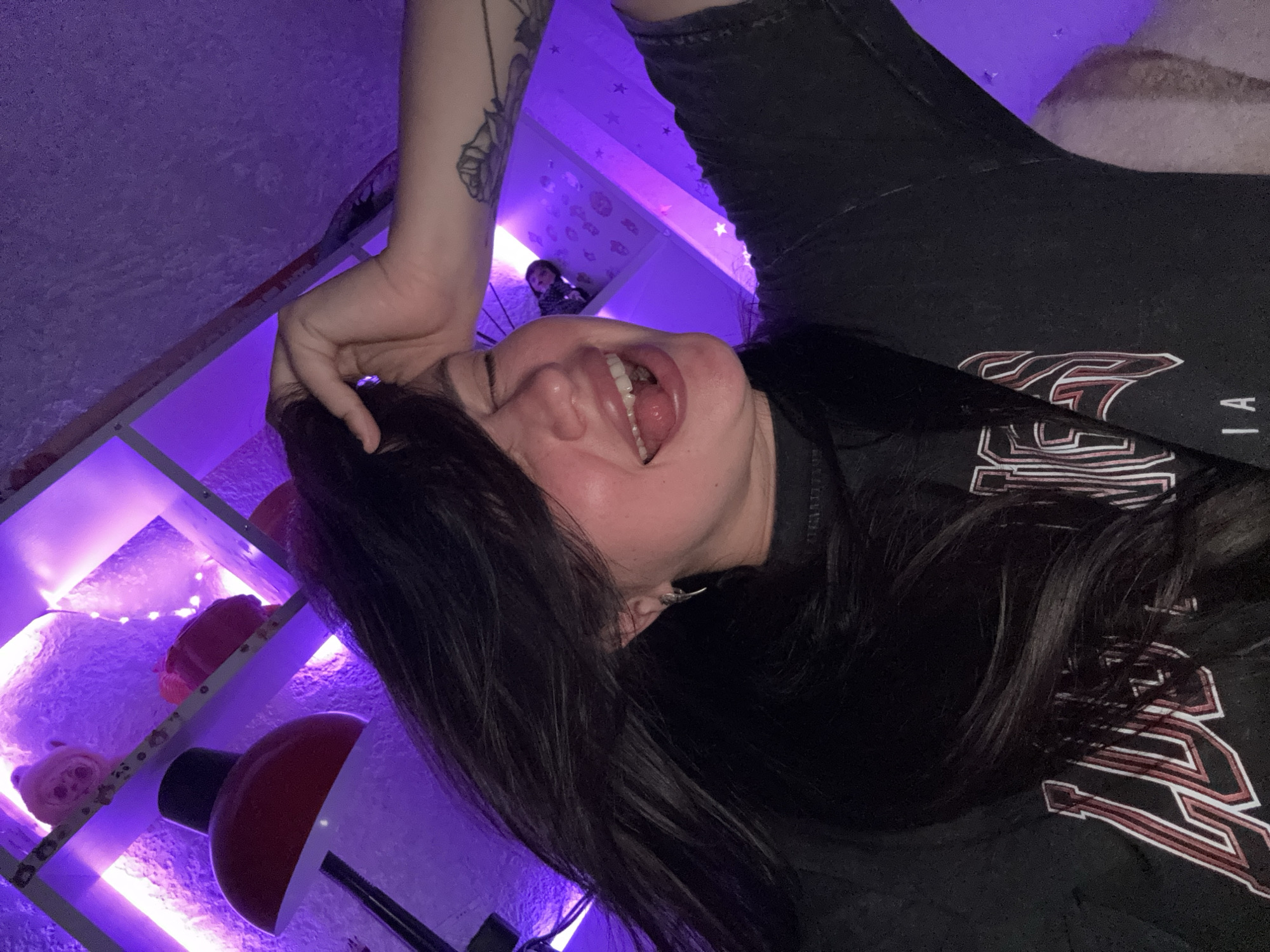A woman with long dark hair is taking a selfie. She is wearing a dark t-shirt with red and white lettering. Her right hand is touching her head, and her left hand is not visible. Her eyes are closed, and her mouth is open, with her tongue sticking out. The background is illuminated by purple light, suggesting a room with decorative lighting. There are shelves with objects on them, including what appear to be stuffed animals and a lamp. The image is underexposed and grainy, typical of a low-light selfie. The location is not identifiable within Tolyatti, Russia, or any other specific geographical location from the visual information provided.