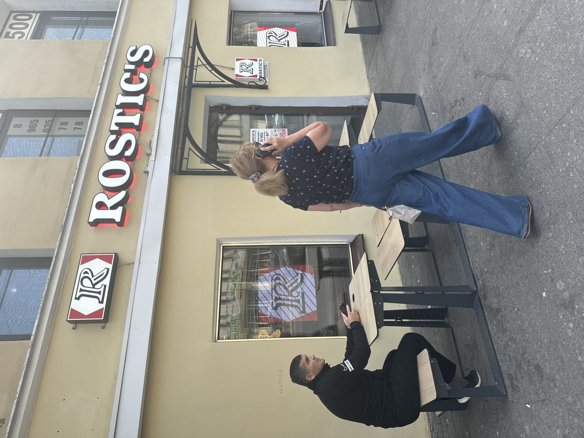 This is an image of the Rostic's restaurant located on Nevsky Prospekt in Saint Petersburg, Russia. A man in a black tracksuit sits at an outdoor table, holding a phone. A woman with blonde hair pulled into a ponytail, wearing a dark floral print shirt and wide-leg blue jeans, walks past the restaurant while talking on her phone. The restaurant has a prominent yellow facade with red and white signage that reads "ROSTIC'S". There are windows displaying a logo with the letter "R". Outdoor seating with tables and benches is present in front of the restaurant. The ground is paved with gray cobblestones.