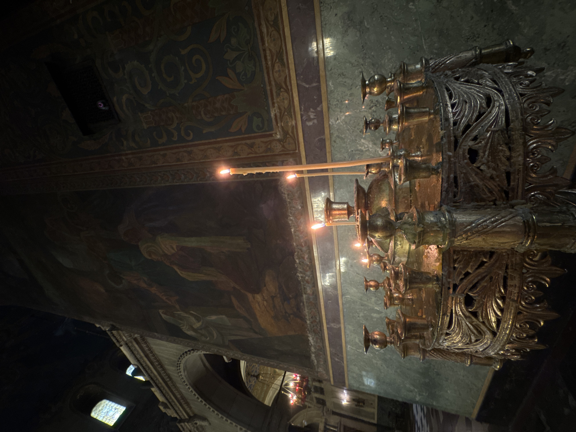 The image depicts the interior of a dimly lit Eastern Orthodox church or cathedral in Sofia, Bulgaria. In the foreground, a heavily ornate, gilded brass candelabrum stands, featuring intricate scrollwork and foliage motifs. Multiple thin, yellow candles are lit on its upper tiers, with significant accumulations of melted wax visible around their bases. The candelabrum is positioned against a wall that exhibits three distinct horizontal sections.
The lowest section of the wall consists of polished greenish-grey marble or stone paneling. Above this is a narrow, horizontal band of purplish-brown material. The largest section, immediately above the band, is adorned with a dark, expansive fresco or mural. This mural depicts several figures, rendered in muted tones of brown, beige, and blue-green, engaged in a scene of religious significance, though specific details are obscured by the low light. Above the mural, the wall transitions to a decorative pattern of stylized blue-green and gold scrolls and floral motifs.
On the left side of the image, a large, stone archway with decorative molding recedes into a darker area of the building. Through this arch, a large, multi-armed chandelier with illuminated red-orange bulbs hangs from a high ceiling. Further up and to the left, two arched windows are visible against a dark overhead structure; one window reveals a pale yellow light, possibly from stained glass. No individuals are present in the scene.