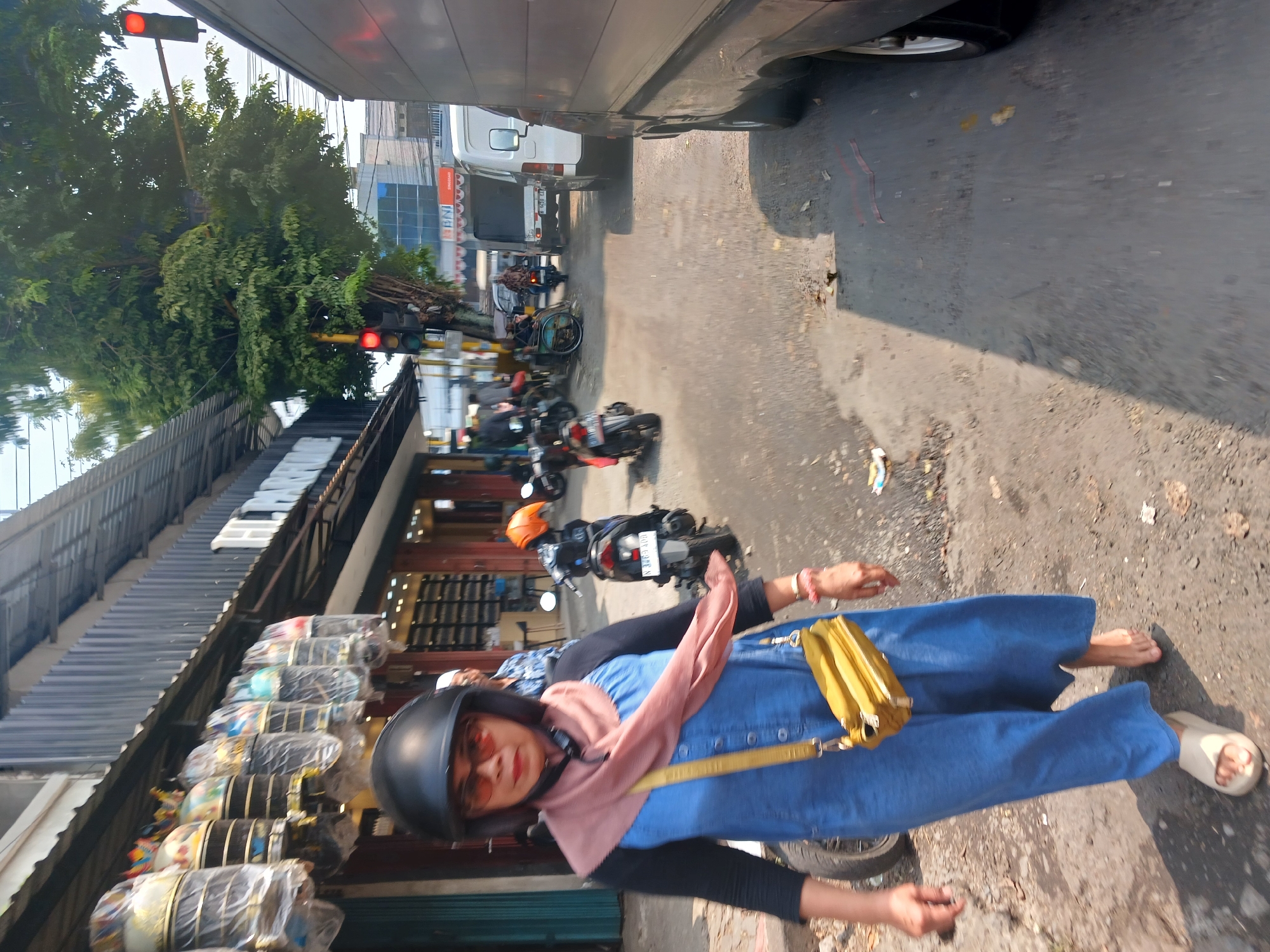 A medium shot captures a woman walking on a street in Singosari, Indonesia. The woman, who is wearing a black helmet, orange-tinted sunglasses, a light pink scarf, and a blue denim jumpsuit, is walking towards the camera. A yellow purse is slung across her body. She is barefoot on her right foot and wearing a light-colored sandal on her left. In the background, several motorcycles are parked, and a building with awnings displaying various items is visible. Traffic lights are present, one of which shows a red signal. A large truck occupies the right side of the frame, obscuring part of the street and adjacent buildings. The street is paved with asphalt and appears somewhat worn, with debris scattered on the ground. Trees line the street, casting shadows, and a bright, sunny sky is visible above.
