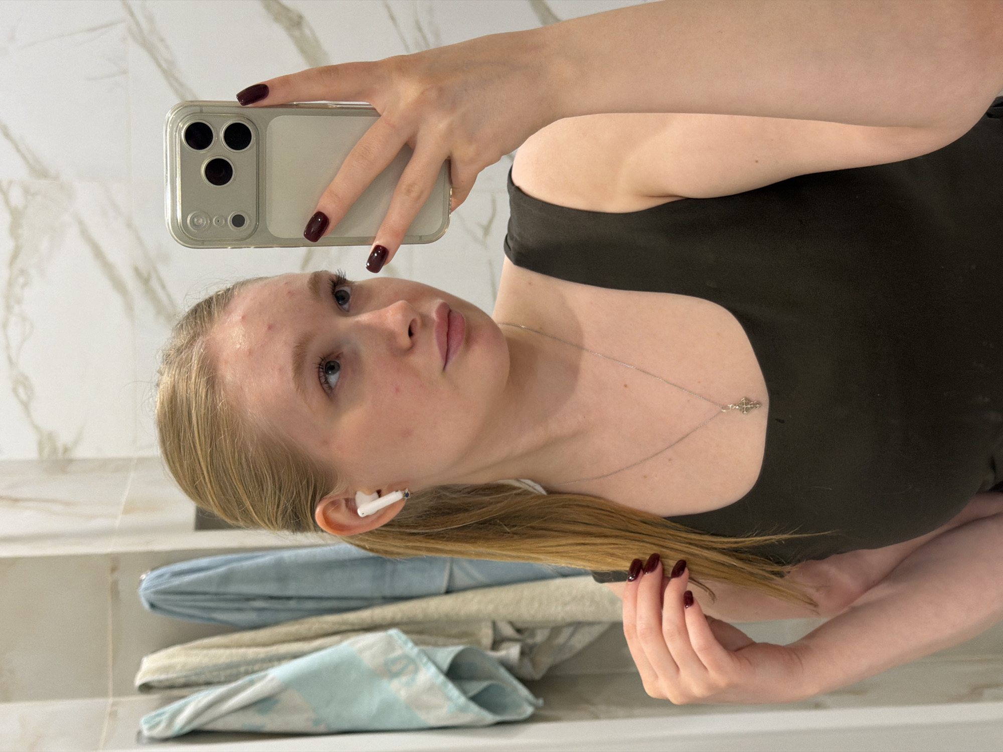 A young woman is taking a selfie in a bathroom. She is holding a smartphone in her left hand, its triple-lens camera visible. She has fair skin, light blonde hair, and blue eyes. She is wearing a black top and a silver necklace with a pendant. An AirPod is visible in her left ear. Her fingernails are painted a dark burgundy color. Background details include marble tiles on the wall and stacked towels on a shelf. The specific location in Rossiyskiy, Russia, cannot be determined from the image.