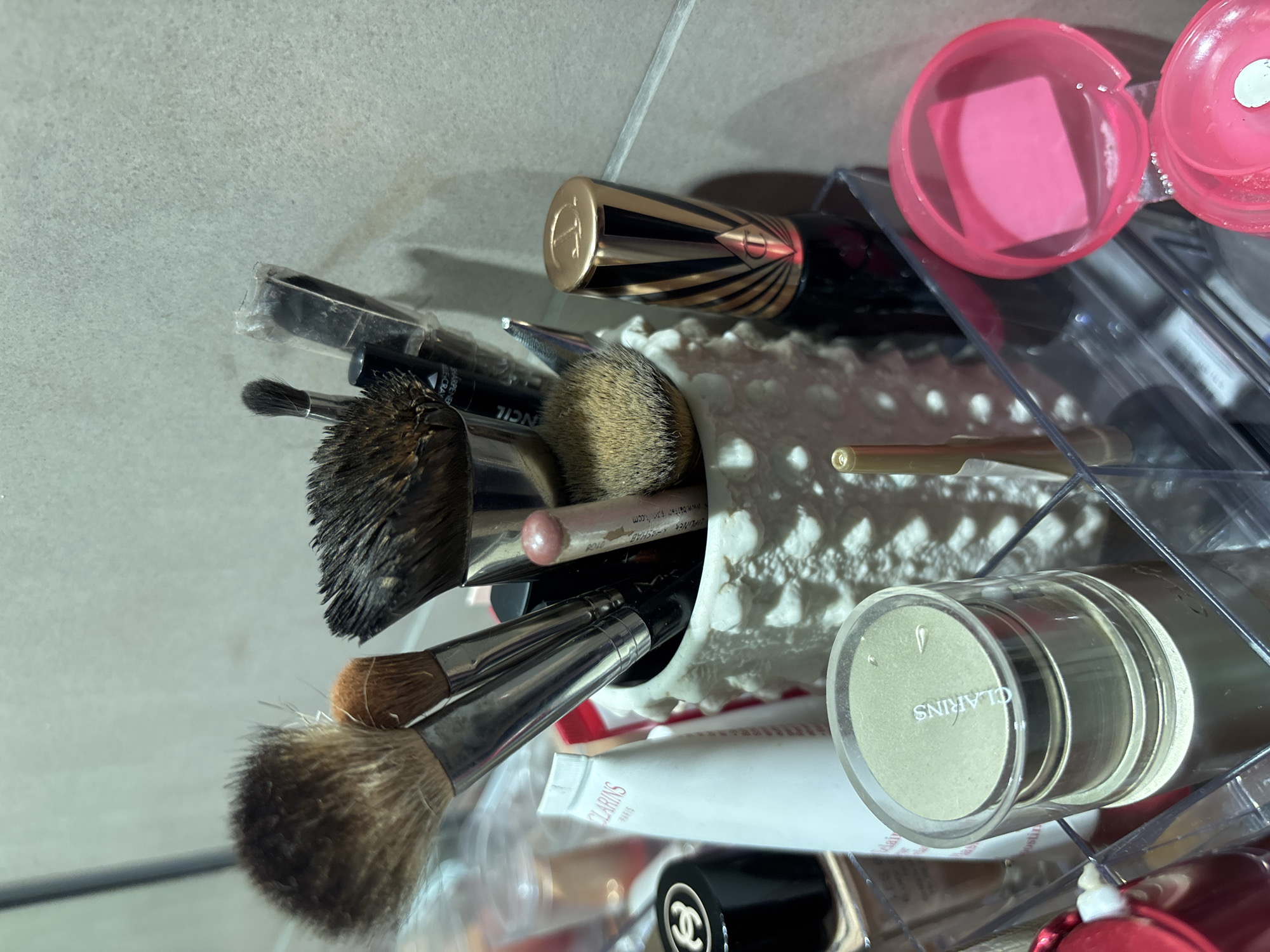 The image shows a collection of makeup products organized in a clear acrylic container and a white ceramic holder. The items include various makeup brushes with natural and synthetic bristles, a tube of Clarins Paris lotion, a black makeup compact with a gold Chanel logo, a Charlotte Tilbury lipstick or similar product with a distinctive black and gold striped packaging and a gold 'T' logo, and a small pink-lidded container with what appears to be a makeup sponge inside. A few other unidentifiable cosmetic items are also visible. The background suggests a bathroom or vanity area with a tiled wall. There are no people or discernible actions in the image. The location of Dubai, United Arab Emirates, is not indicated by any visual cues in the provided image.