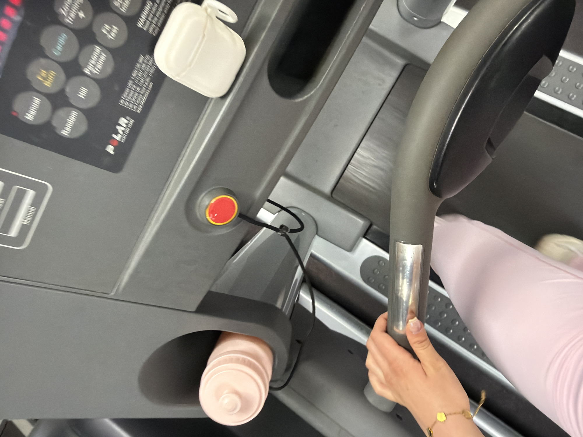 The image shows a close-up view of a treadmill in what appears to be a gym setting. The treadmill's control panel is visible, featuring buttons labeled with workout programs. A red emergency stop button is also present. A white object, possibly headphones, is positioned near the control panel.
A person is gripping one of the treadmill's handles. Only the arm and hand are visible, adorned with a gold bracelet. The person appears to be wearing light pink athletic wear. The treadmill belt is partially visible, along with the person's legs, suggesting active use. A pink water bottle is placed in the treadmill's holder.
Given the description and the presence of a gym, it is not possible to determine the location in the city of Sofia, Bulgaria.