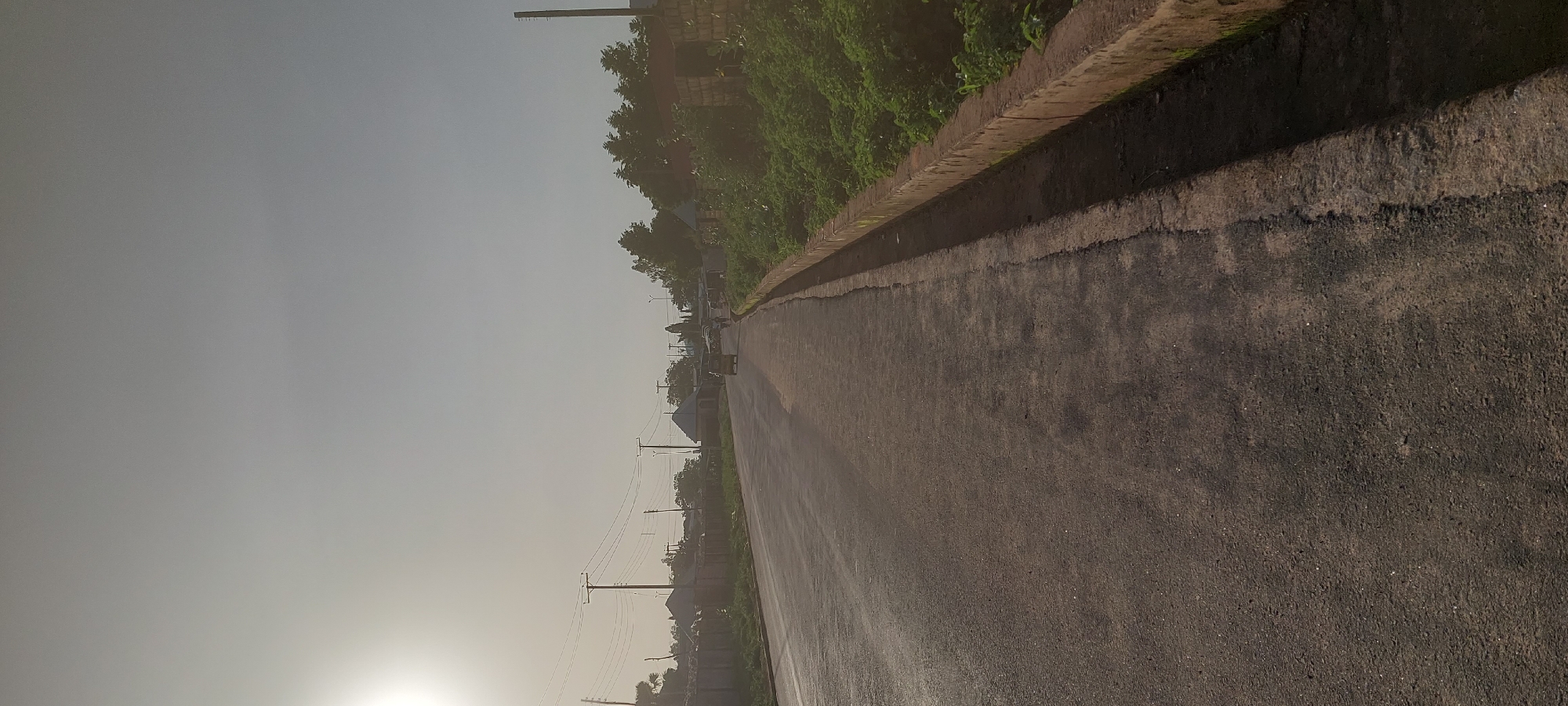This is a vertical, eye-level shot of a paved road in Jalingo, Nigeria, under a bright, hazy sky. The sun is visible in the upper left corner, casting a strong glare and a soft, diffused light across the scene. The road itself is dark asphalt and appears wet or recently rained on, with reflections visible on its surface. To the right of the road is a concrete gutter with a rough, textured edge, and beyond that, a dense growth of green vegetation.
Along the left side of the road, there is a low, rough-textured wall made of stones and cement. Several utility poles with wires are scattered along the landscape to the left and in the distance. In the background, various structures with pitched roofs and some open-sided market stalls or shelters are visible. A few vehicles, including what appears to be a yellow tuk-tuk and a white van, are traveling down the road in the distance. A solitary person is walking on the right side of the road. The overall impression is a street scene in a developing urban or semi-urban area on a bright, overcast day.