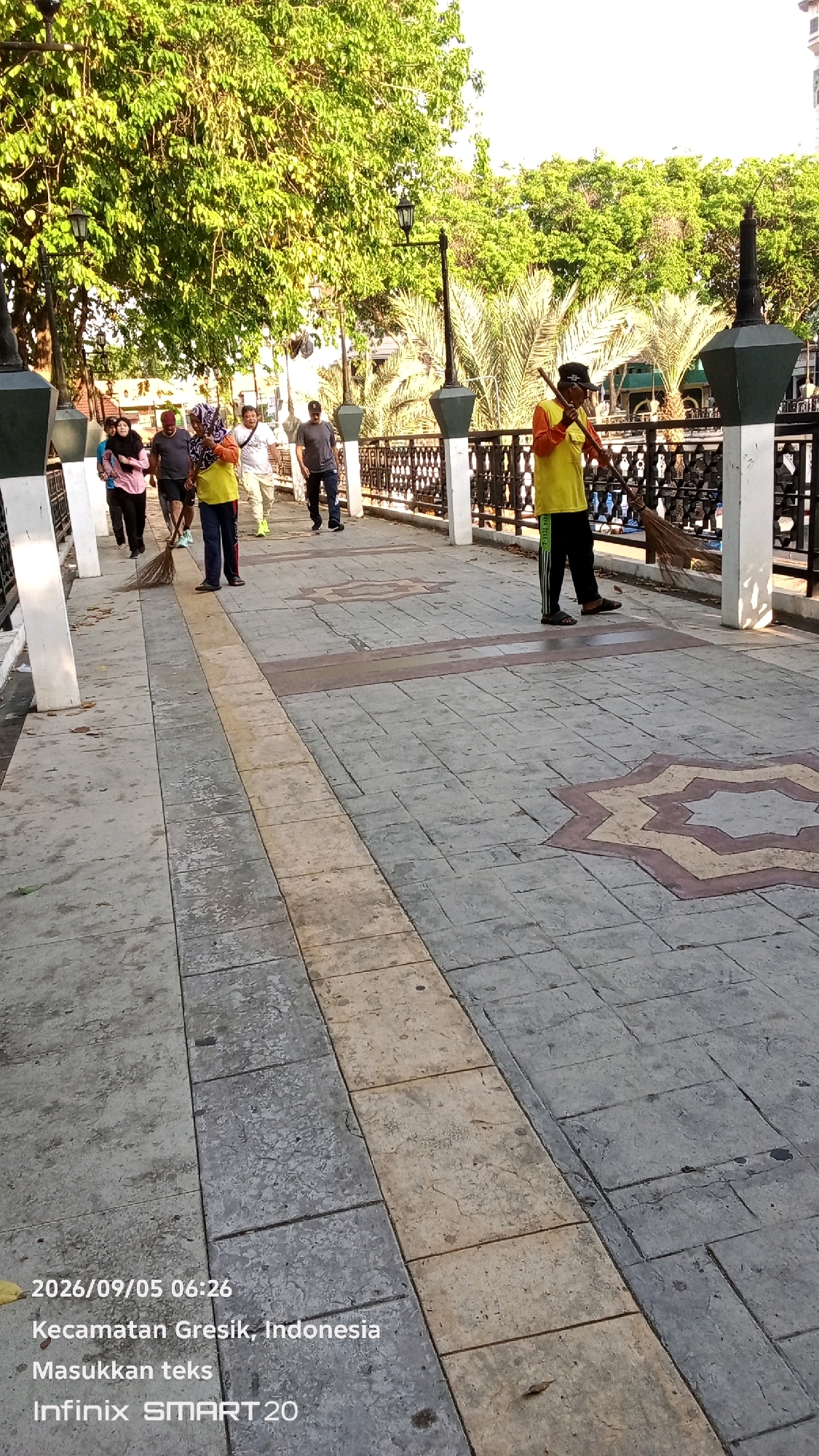 A street sweeper in a yellow vest and black pants is sweeping a patterned concrete walkway with a broom. Several other people, including some wearing headscarves, are walking on the walkway in the background. The walkway is lined with decorative lampposts and a black metal fence. Lush green trees and palm trees are visible in the background, suggesting an outdoor setting in a city park or public space. The timestamp on the image indicates it was taken on September 5, 2026, at 06:26 in Gresik, Indonesia.