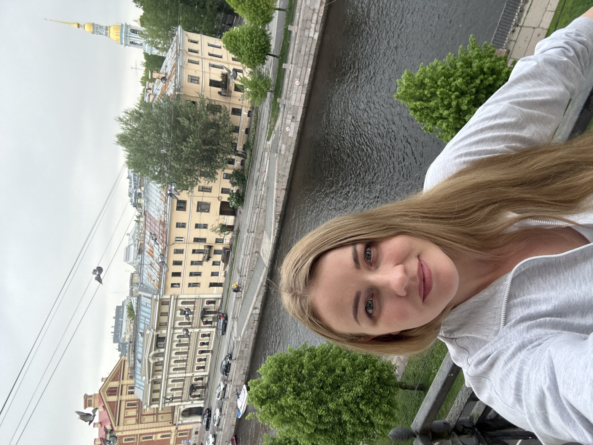 A woman poses for a selfie-style photograph in Saint Petersburg, Russia. She is positioned on a bridge, overlooking a canal, with buildings visible in the background. The Pavlovsky Regiment Life Guards Cathedral is seen in the background. The day appears to be overcast.