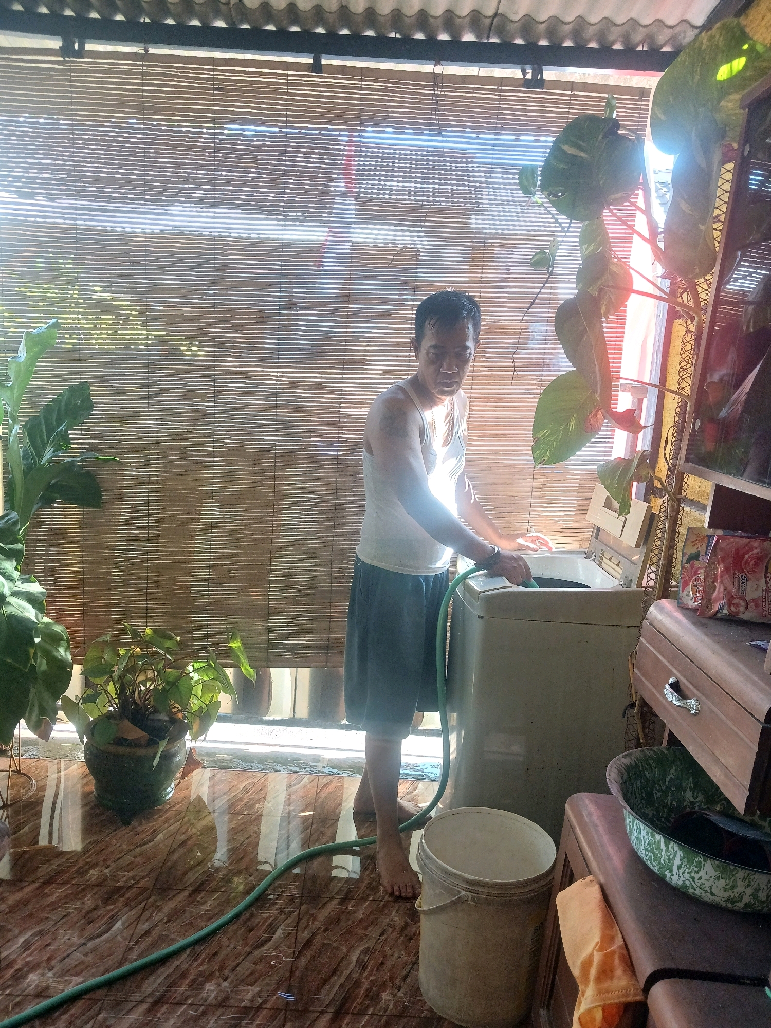 In Banyuwangi, Indonesia, a man in a white tank top and blue shorts stands in a dimly lit, rustic room, preparing to do laundry. He is holding a green hose, which appears to be connected to a top-loading washing machine. Sunlight streams through a bamboo blind, casting long shadows across the tiled floor and a large potted plant. To the right of the washing machine, a wooden cabinet with a drawer and a green and white patterned basin sits on a stand. A yellow cloth is draped over the stand. A white bucket and another potted plant are visible on the floor. The scene suggests a domestic setting with limited modern amenities.